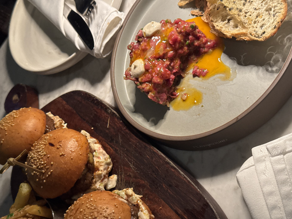 A plate presents raw beef tartare with a vibrant egg yolk, alongside a slice of toasted bread sprinkled with seeds. Nearby, a wooden board displays mini burgers, their sesame seed buns filled with meat and a creamy topping. White plates, napkins, silverware and fries accompany the meal on a marble surface.