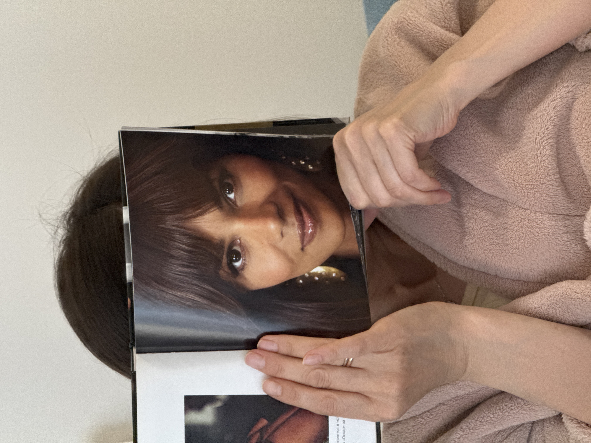 A woman wearing a fuzzy, peach-colored robe is holding a magazine in front of her face. The magazine features a portrait of a woman with brown hair and brown eyes. The scene is indoors. The location is not evident in this image and cannot be inferred.