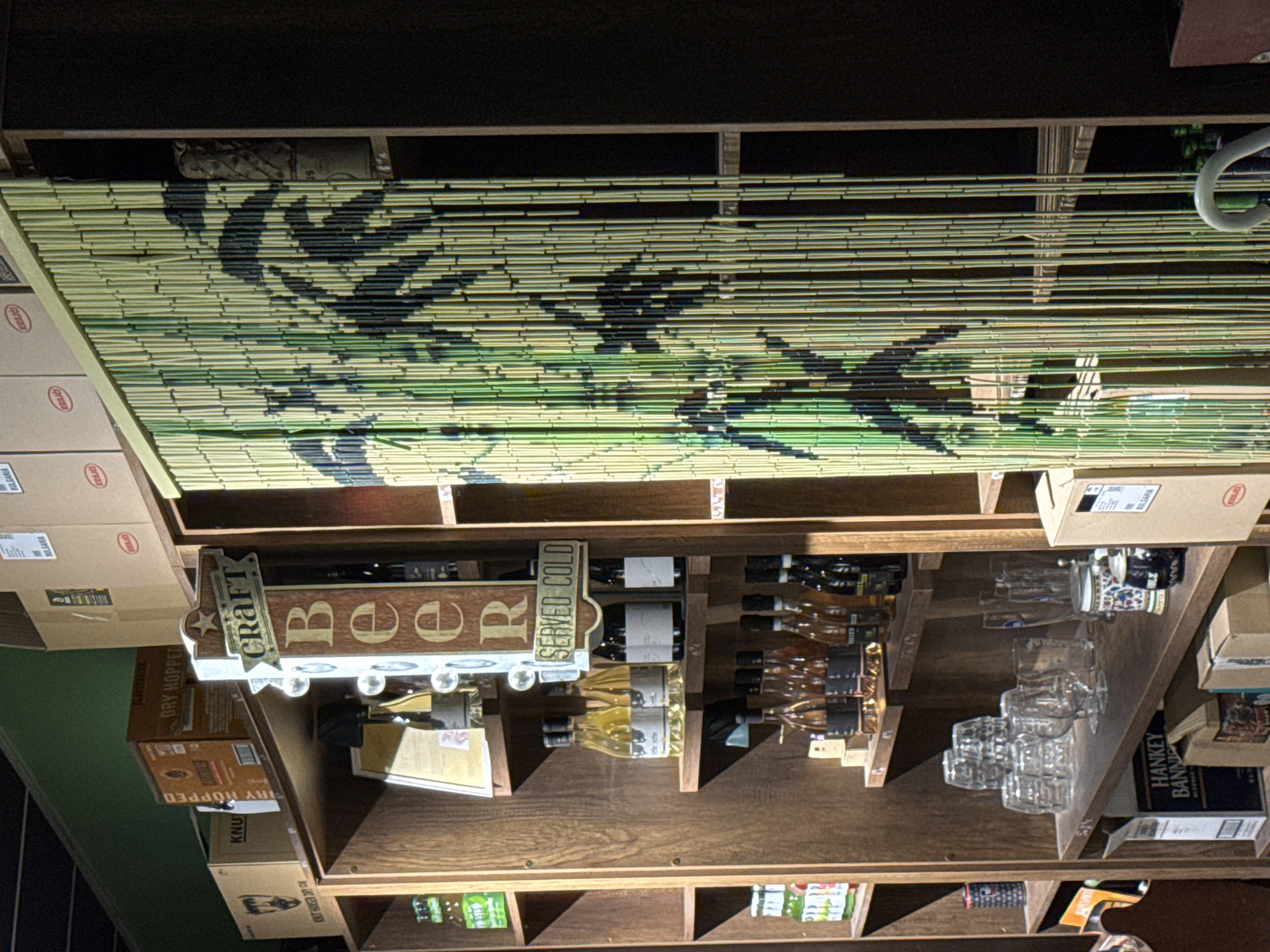 The image shows the interior of what appears to be a bar or restaurant. The primary focus is on a wooden shelf stocked with various items. There are rows of bottles, including wine, and glasses arranged on the shelf. Signage reads "CRAFT Beer SERVED COLD." There are boxes labeled with brands such as "DRY HOPPED," "KNUT," and "HANKEY BANNI," as well as the word "BULGARIA." The environment suggests a place that serves alcoholic beverages. Above the shelf, there's a bamboo-like hanging divider with a dark pattern on it. The setting and markings might indicate it is somewhere in Bulgaria, likely Veliko Tarnovo, where there is an ale house.