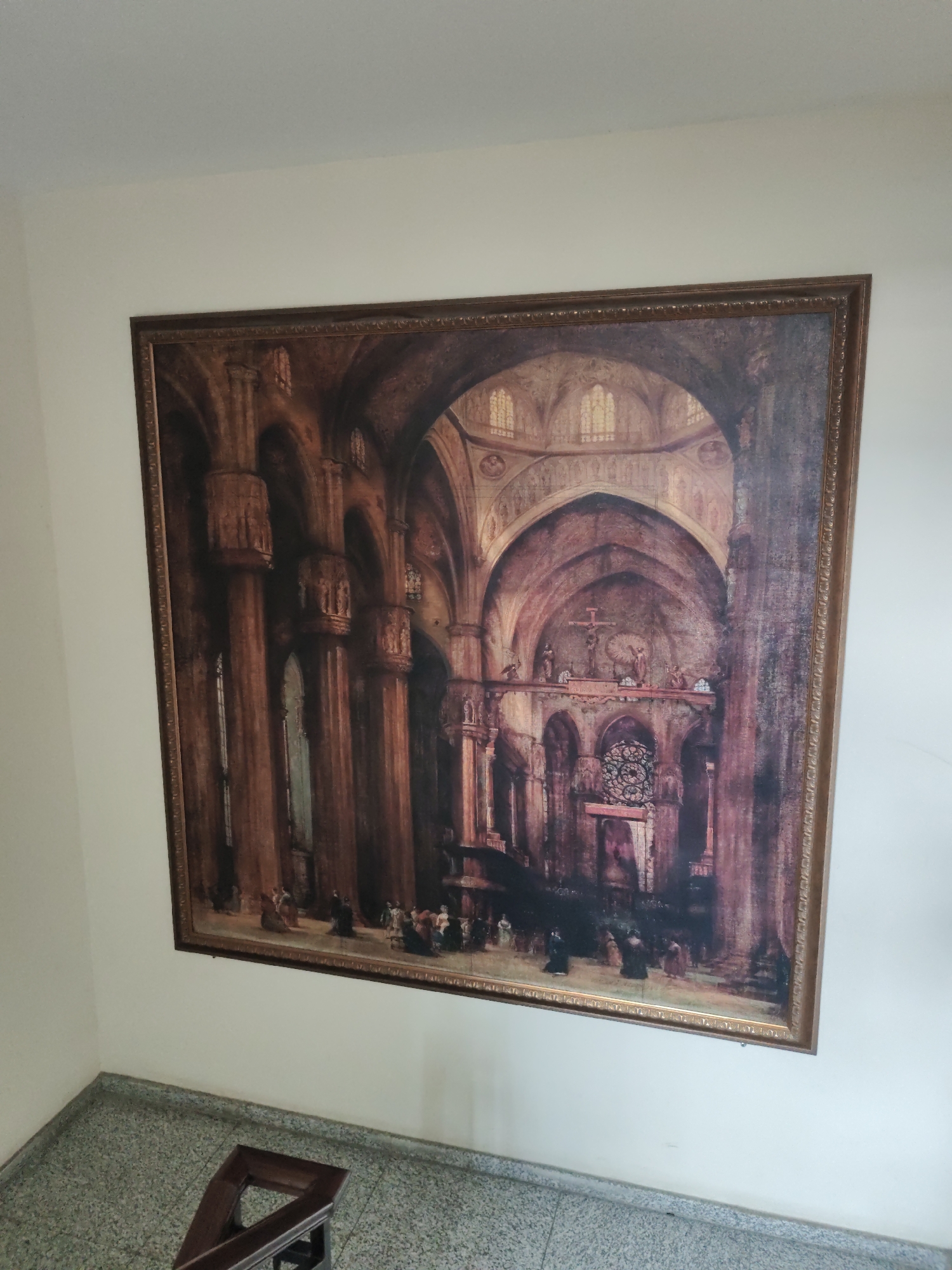 A framed oil painting depicts the interior of a grand cathedral, rendered in dark, rich tones of brown and red, with shafts of light filtering through arched windows. The architecture features massive columns, soaring arches, and intricate stonework, including a prominent rose window above an altar with a crucifix. Small figures, appearing to be monks or clergy in dark robes, are scattered throughout the nave, engaged in prayer or contemplation. The painting is hung on a plain, cream-colored wall. Below the painting, a speckled grey floor leads to a dark wooden railing, part of a staircase. The image was captured in the city of Aihui, China.