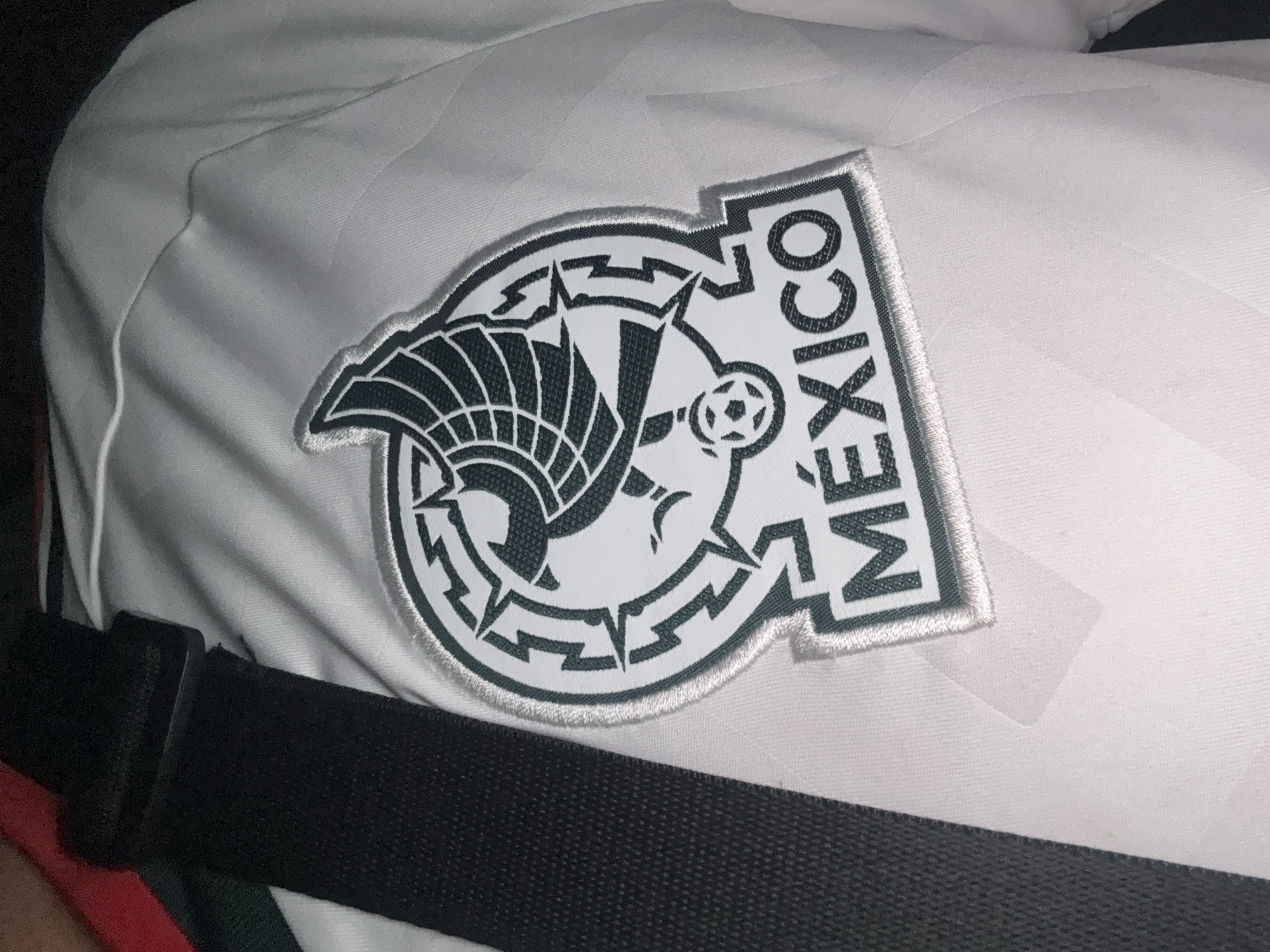 The image shows a close-up of a white soccer jersey with a dark green and white emblem on the chest. The emblem features an eagle with its wings spread, perched on a cactus, and the word "MÉXICO" below. A black strap runs diagonally across the left side of the jersey. The background is blurred and out of focus, but appears to be indoors. There is no specific information or visual cues to indicate the location is in Mexico City, Mexico. The image is a static, unposed shot of the jersey.