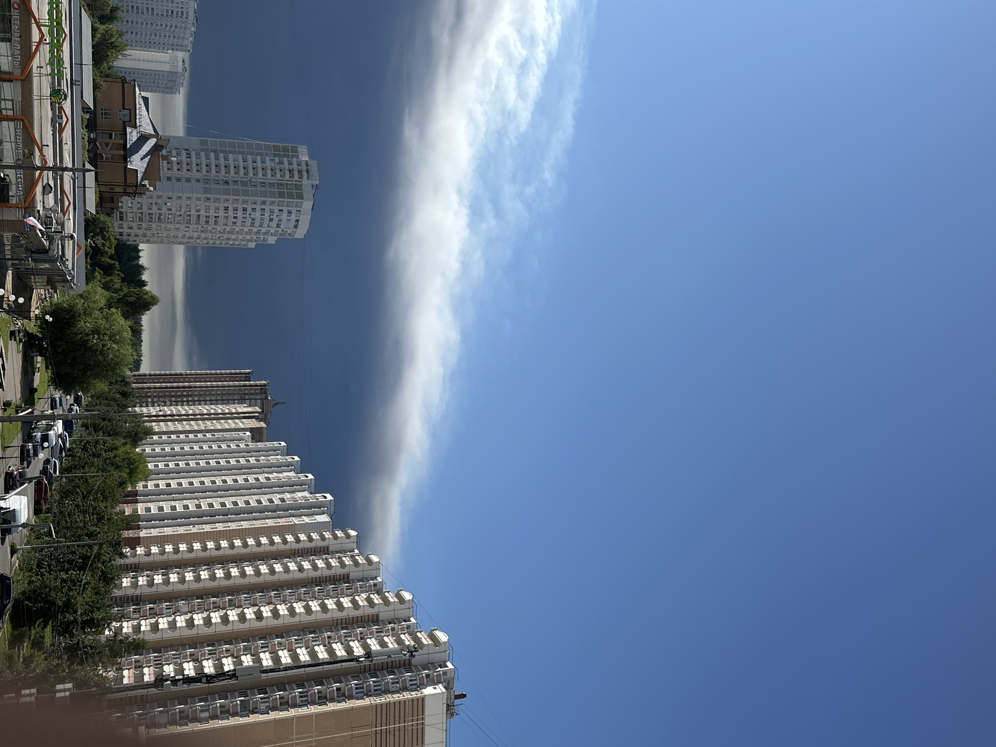 The image shows a panoramic view of a residential area in Podolsk, Russia, under a dramatic sky. In the foreground, a row of tall, multi-story apartment buildings with white facades and many balconies stretches across the frame, suggesting a densely populated urban environment. The buildings are modern in design, with a repeating pattern of windows and balconies. Behind these buildings, a few other high-rise structures are visible, one of which is a distinct, slender white tower. A dark, ominous band of clouds dominates the middle ground of the sky, creating a stark contrast with the clear, bright blue sky above. This cloud formation appears to be a weather front, possibly indicating an approaching storm. Below the cloud band, a line of lush green trees partially obscures some of the lower buildings and a road where several cars are parked or moving. In the lower left corner, the tops of some commercial buildings with visible signage are present. The overall impression is one of urban development set against a backdrop of changing weather.