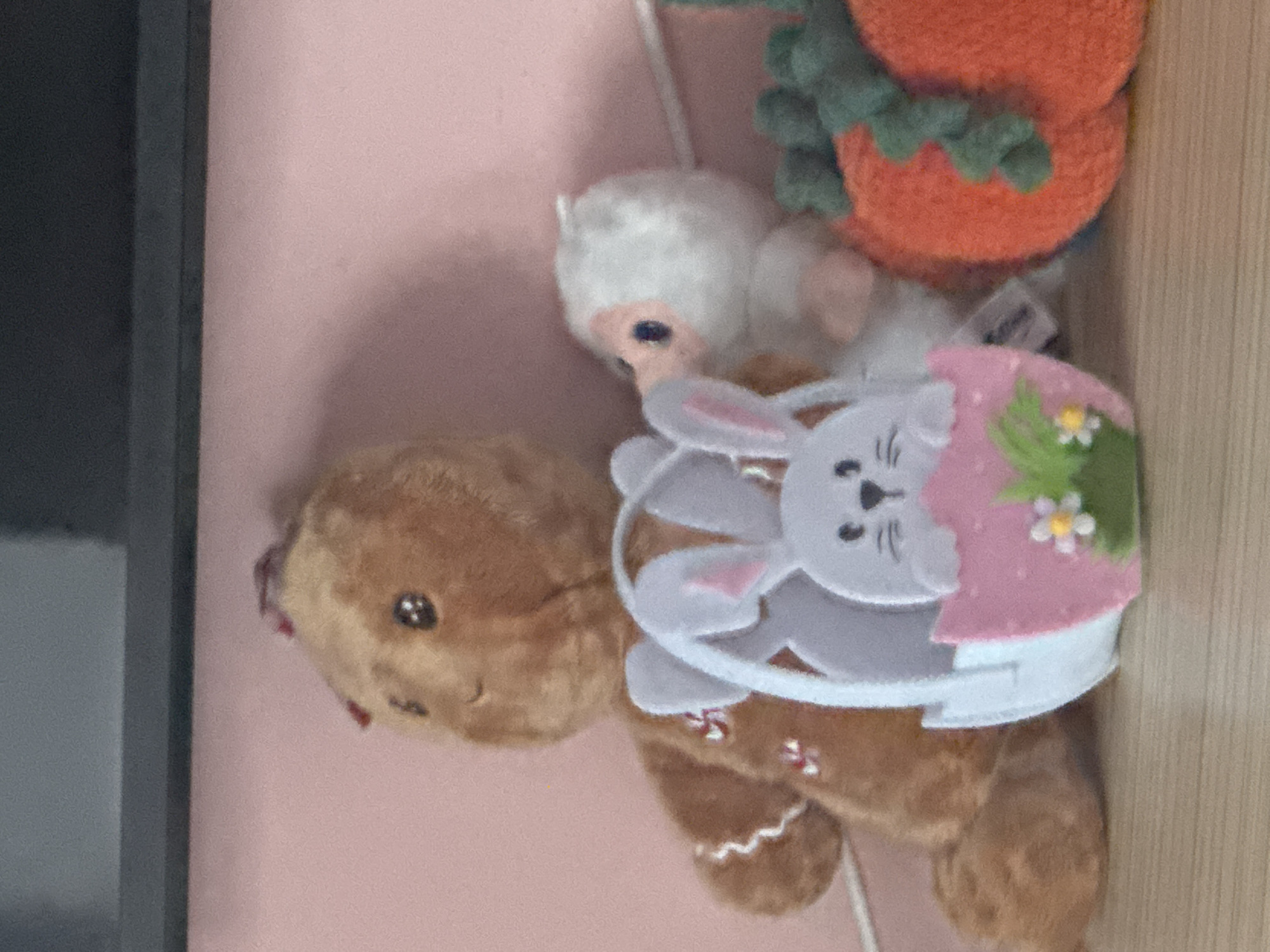 An indoor scene displays a collection of plush toys and decorative objects arranged on a light-colored wooden surface. The background consists of a plain, light pink wall, with a dark gray or black horizontal structure (possibly a shelf or furniture top) visible across the upper portion.
From left to right, the prominent objects are:
1. A brown plush gingerbread figure, sitting upright, with black button eyes, a stitched smile, white zig-zag "icing" patterns on its limbs, and small red and white stitched accents on its torso. A red ribbon is partially visible on its head.
2. In front of the gingerbread figure and slightly to its right, a white felt or fabric decorative basket. The basket features cut-out rabbit ears on its top rim and a white rabbit head (with black eyes and nose) peering from a pink egg-shaped opening. The front of the egg-shape is adorned with a green patch of "grass" and a small white flower with a yellow center.
3. Behind the basket and to the right of the gingerbread figure, a small, white plush toy with a light pink face/beak area and blue eyes.
4. To the far right, partially visible, is an orange plush decoration resembling a segmented pumpkin or squash, with dark green stem/leaf elements.
The objects are positioned on a light brown surface exhibiting a faint horizontal wood grain. The lighting is even and ambient, without harsh shadows. The location depicted is undetermined; no city or specific address can be ascertained from the visual information provided.