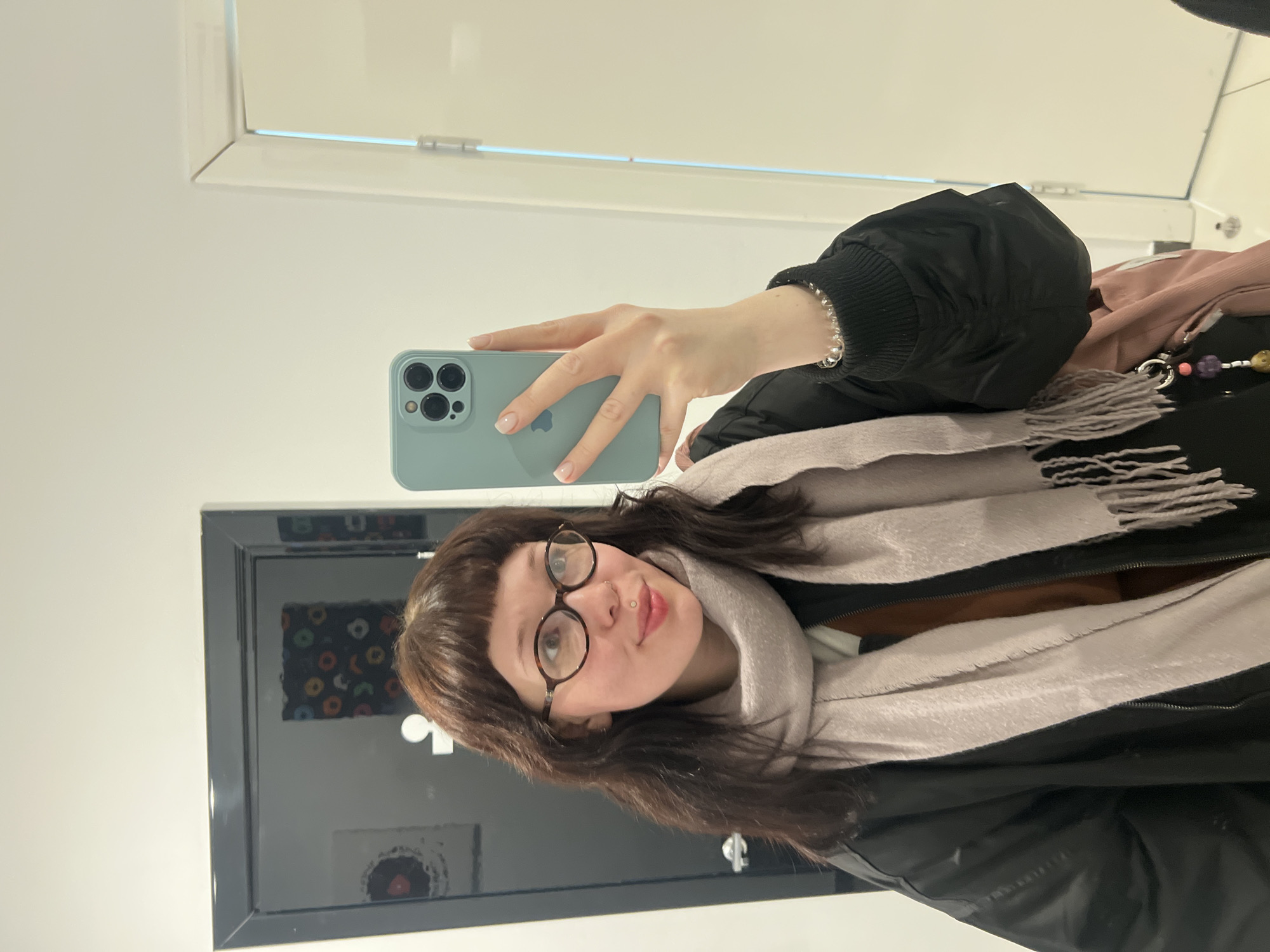 A young woman is captured in a mirror selfie, holding a light blue/mint green iPhone with three visible camera lenses and an Apple logo on its back. The image is rotated 90 degrees clockwise.
The woman has medium-length dark brown hair with bangs and is wearing round, dark-framed glasses. She has a subtle smile and a labret piercing below her lower lip. She is dressed in a black jacket, possibly a puffer or bomber style, with ribbed cuffs, and a light pink or beige scarf with fringed edges wrapped around her neck. A brown garment is visible underneath her jacket. On her right wrist, she wears a silver beaded bracelet. Part of a bag strap with colorful beaded charms is visible near her shoulder.
The background shows a simple, light-colored (off-white/cream) wall. The large, dark-framed mirror she is posing in reflects a part of the room behind her, revealing abstract art or patterned circular items in various colors on a darker surface, possibly wall decor or framed pictures. A white door or window frame with a faint blue line is visible in the upper right portion of the reflected area. The setting appears to be indoors, well-lit, and suggesting a modern interior. The jacket and scarf imply a cooler temperature. No explicit text is visible other than the Apple logo on the phone. The general location context is Chaikovsky, Russia.