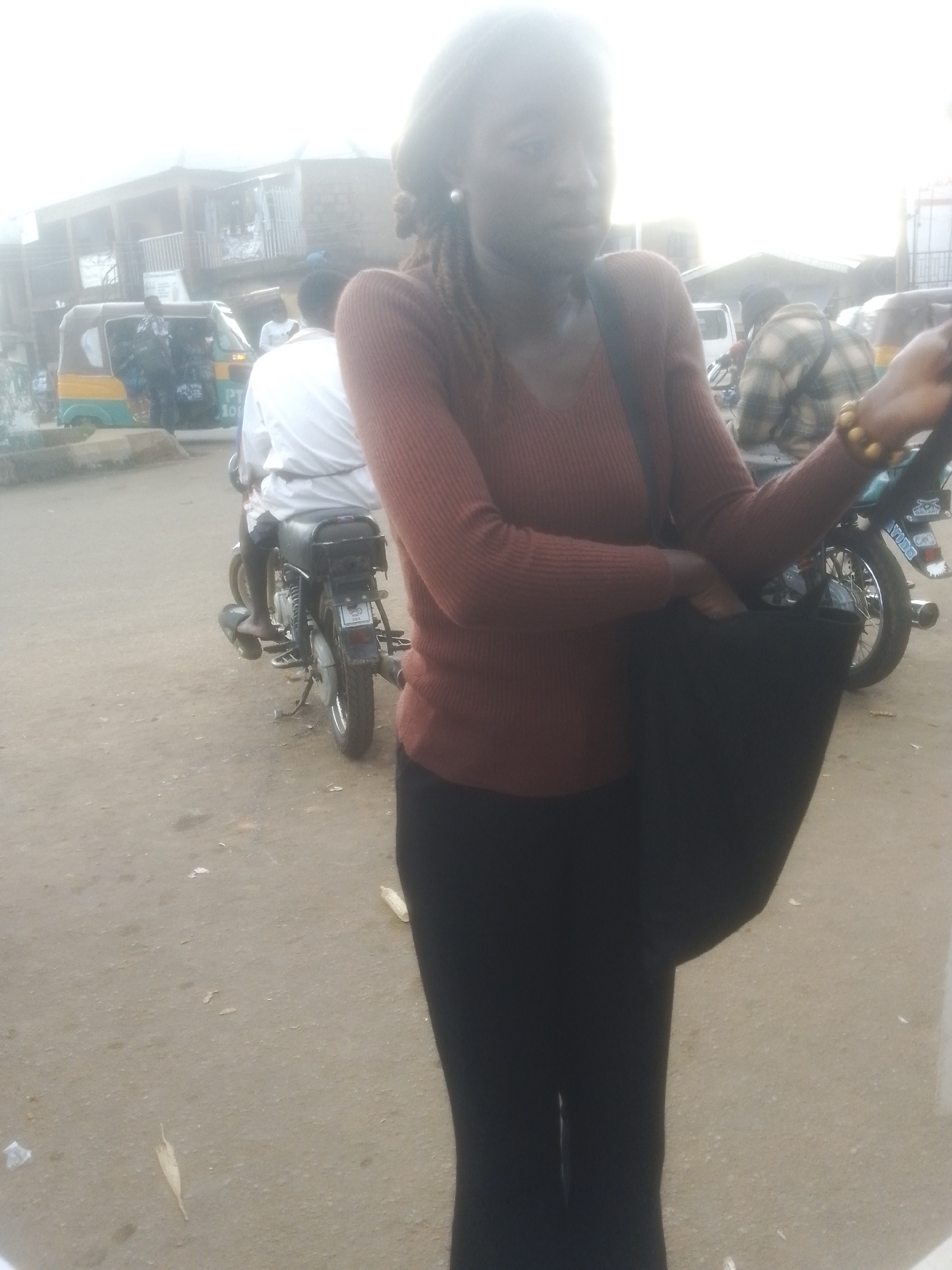 A young African woman, wearing a rust-colored knitted sweater and black pants, stands in the foreground, holding a large black tote bag. She has dark skin, dark hair, and appears to be wearing an earring. A beaded bracelet adorns her right wrist. In the middle ground, a person wearing a white shirt sits on a motorcycle. Behind them, a yellow and green auto-rickshaw is parked. In the background, buildings and other vehicles are visible, suggesting an urban environment, possibly a market or transportation hub. The ground is paved with asphalt or dirt, and there are scattered pieces of debris. The overall lighting suggests an overcast or hazy day. The scene is consistent with an outdoor street setting in Gwong, Nigeria.