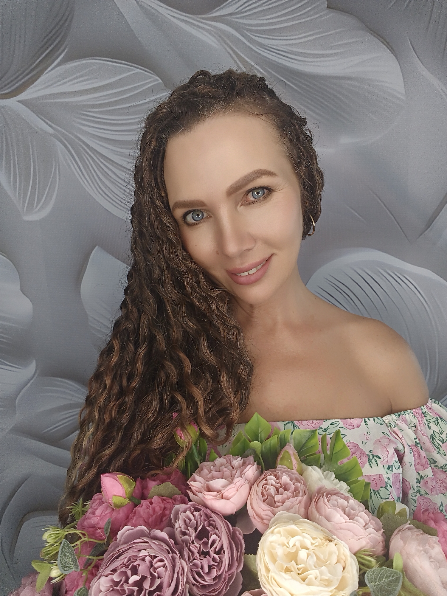 A medium shot shows a woman with curly brown hair and blue eyes holding a bouquet of pink and cream flowers. She is wearing an off-the-shoulder dress with a floral print. The background is a textured gray wall with a light floral pattern. The woman is smiling and looking at the camera. There are no other people or animals visible in the image. The scene appears to be indoors, possibly in a studio or home setting. The lighting is soft and even, highlighting the woman's features and the flowers.