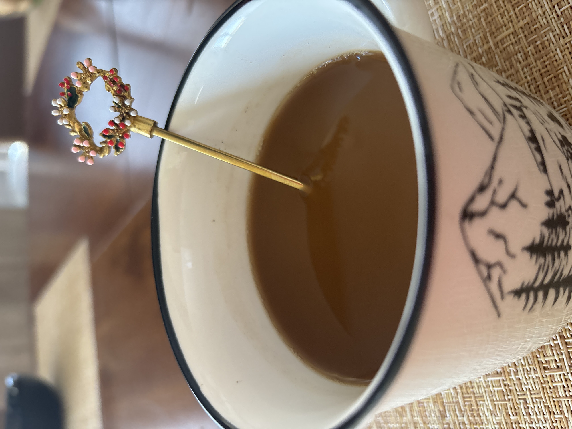 A close-up, eye-level shot shows a mug of coffee with a decorative stirring stick. The mug is white with a black rim and a black silhouette of mountains and pine trees. The coffee is light brown, and a thin gold stirring stick with a small, colorful wreath-like ornament is partially submerged in it. The mug sits on a light brown woven placemat on a wooden table. In the blurred background, a dark bowl is visible. The scene appears to be indoors, possibly in a home or cafe. There are no people visible in the image. The location is not specified as Sochi, Russia, nor is there any information to suggest this is the case.