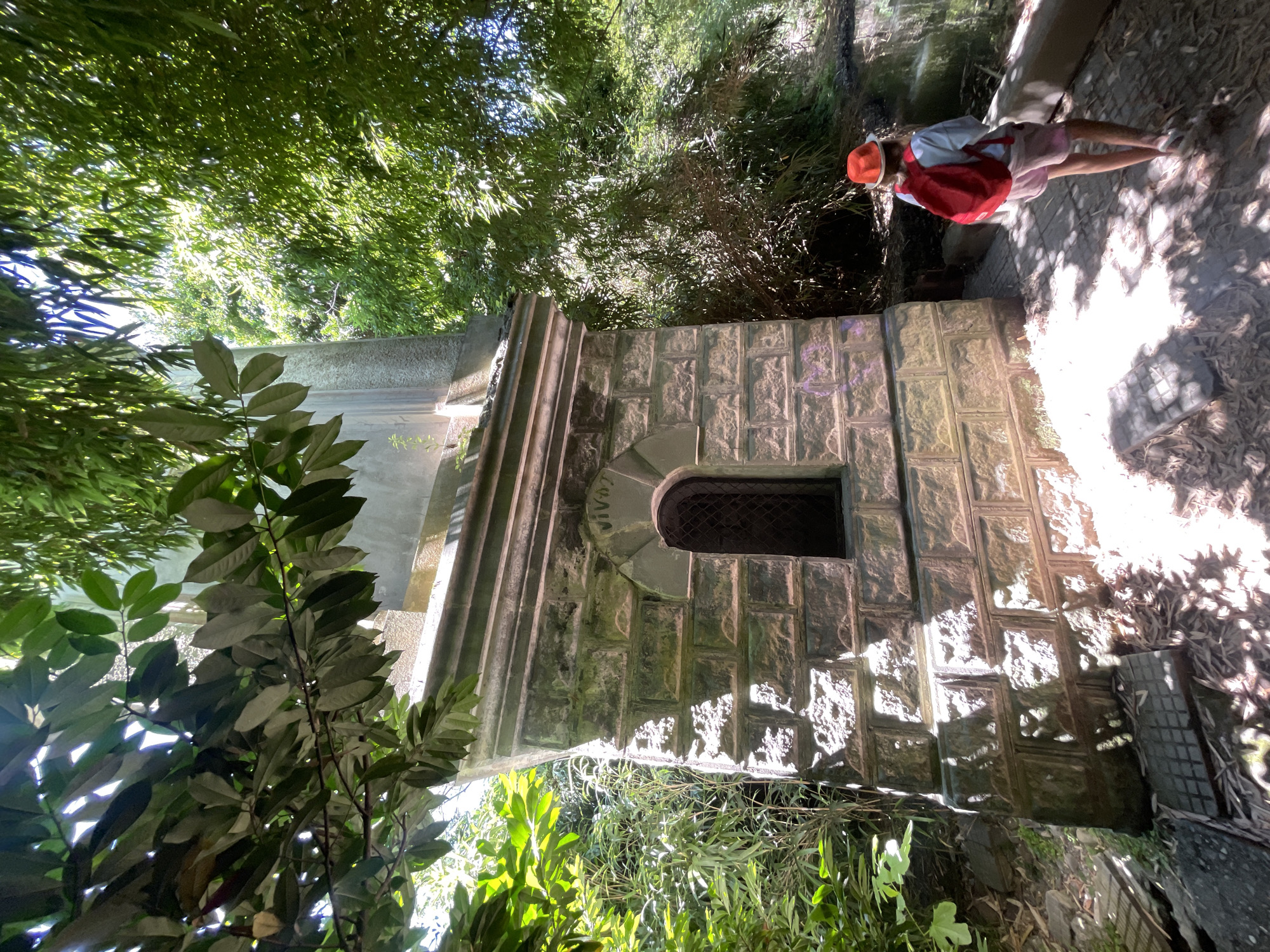 A young person wearing a red backpack, a white shirt, and pink shorts walks away from the camera down a path next to a stone structure. The person also wears an orange hat. The stone structure has a small arched window with a grid. The structure is surrounded by lush green foliage, and sunlight filters through the leaves, creating dappled shadows on the ground. The scene appears to be in a park or natural area.