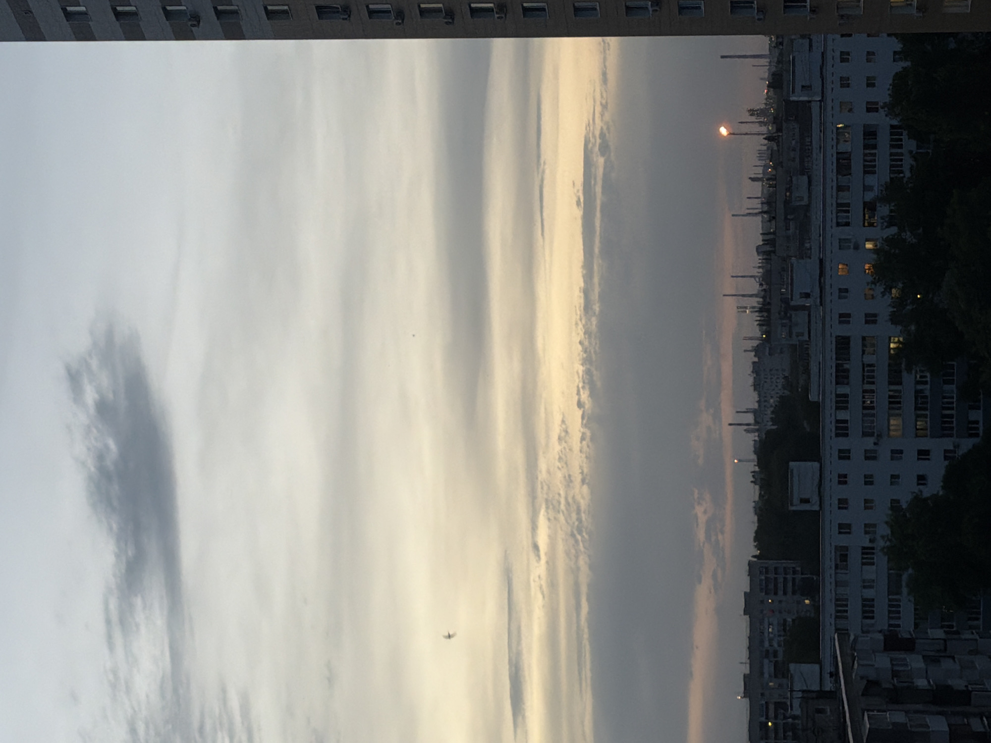 The image presents a cityscape under an overcast sky. In the foreground, the top of a building is visible, framing the view. The central area is dominated by a cloudy sky with varying shades of gray and a hint of yellow. A small plane is visible amidst the clouds. In the background, there are several buildings, with a cluster of industrial chimneys and a street lamp standing out. The overall scene suggests an urban environment during the late evening or early morning.