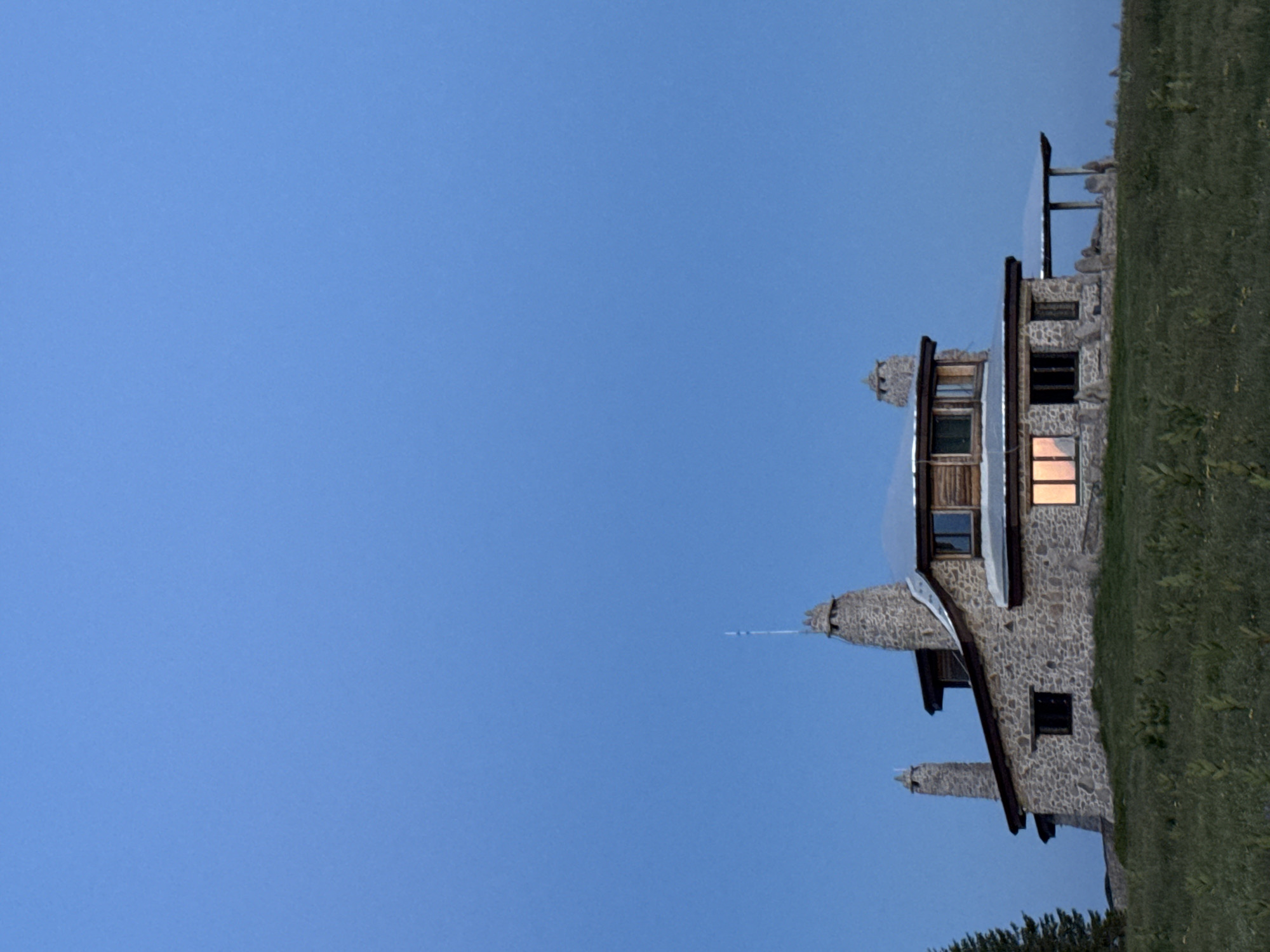 A stone house with multiple chimneys sits on a grassy hill under a clear blue sky. The house has a wooden roof and several windows. One window glows with warm light from inside. A covered porch with stone pillars extends from the side of the house. A single evergreen tree is visible to the left of the house. The image appears to be taken at dusk or dawn, as the sky is a deep, even blue and the light is soft. There is no indication of the city of Gela, Bulgaria, or any people in the image.