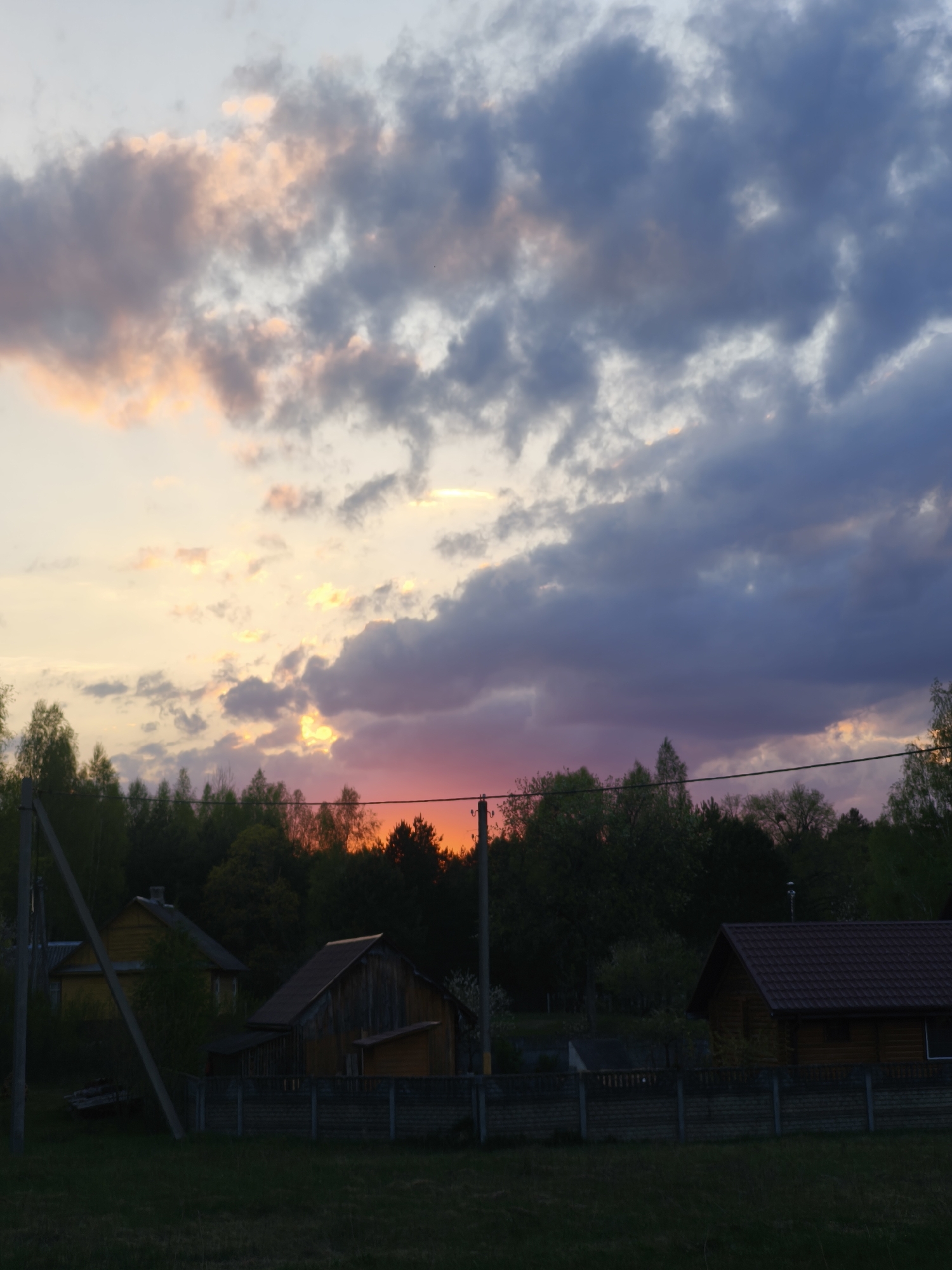 The image depicts a rural scene at dusk in Парэчча, Belarus. The sky is filled with dark, cloudy formations with patches of orange and pink light. Silhouetted trees form a line across the mid-ground, with the horizon illuminated by the setting sun, which appears red. Three houses are visible in the lower part of the frame behind a low concrete fence. Power lines and poles cut across the sky and landscape. The foreground consists of a grassy field.