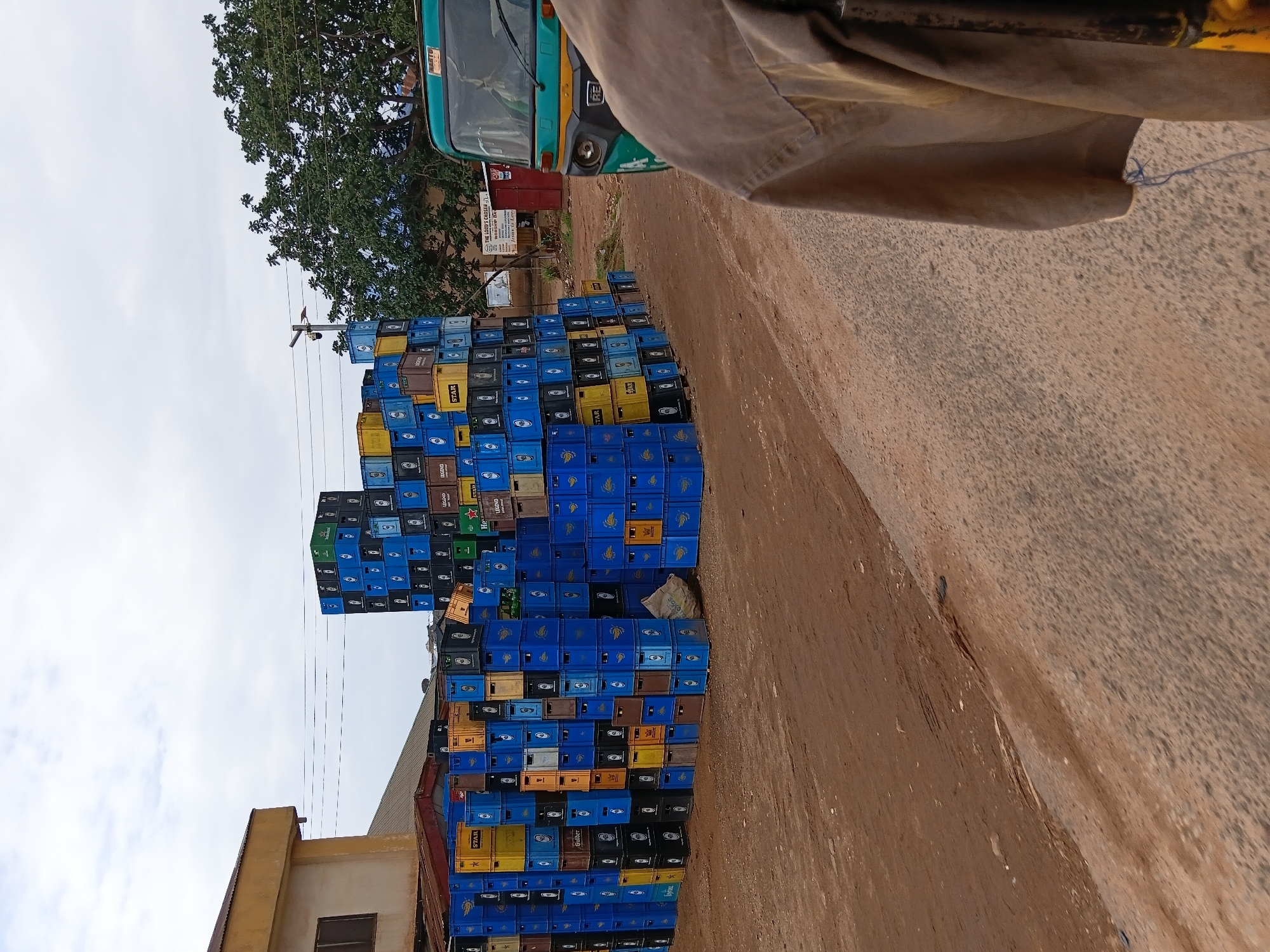 A large stack of blue, black, and yellow beer crates, some labeled "STAR," "Gulder," and "LEGEND," are piled precariously high on a dirt road in Gwong, Nigeria. The stack is adjacent to a low building with a yellow facade. Overhead power lines run near the top of the stack. A green and yellow auto rickshaw is partially visible in the background. Part of a person wearing a tan shirt is visible on the right side of the frame. A sign reading "THE LORD'S CHOSEN WORSHIP DAY" is visible in the background, indicating a religious institution.