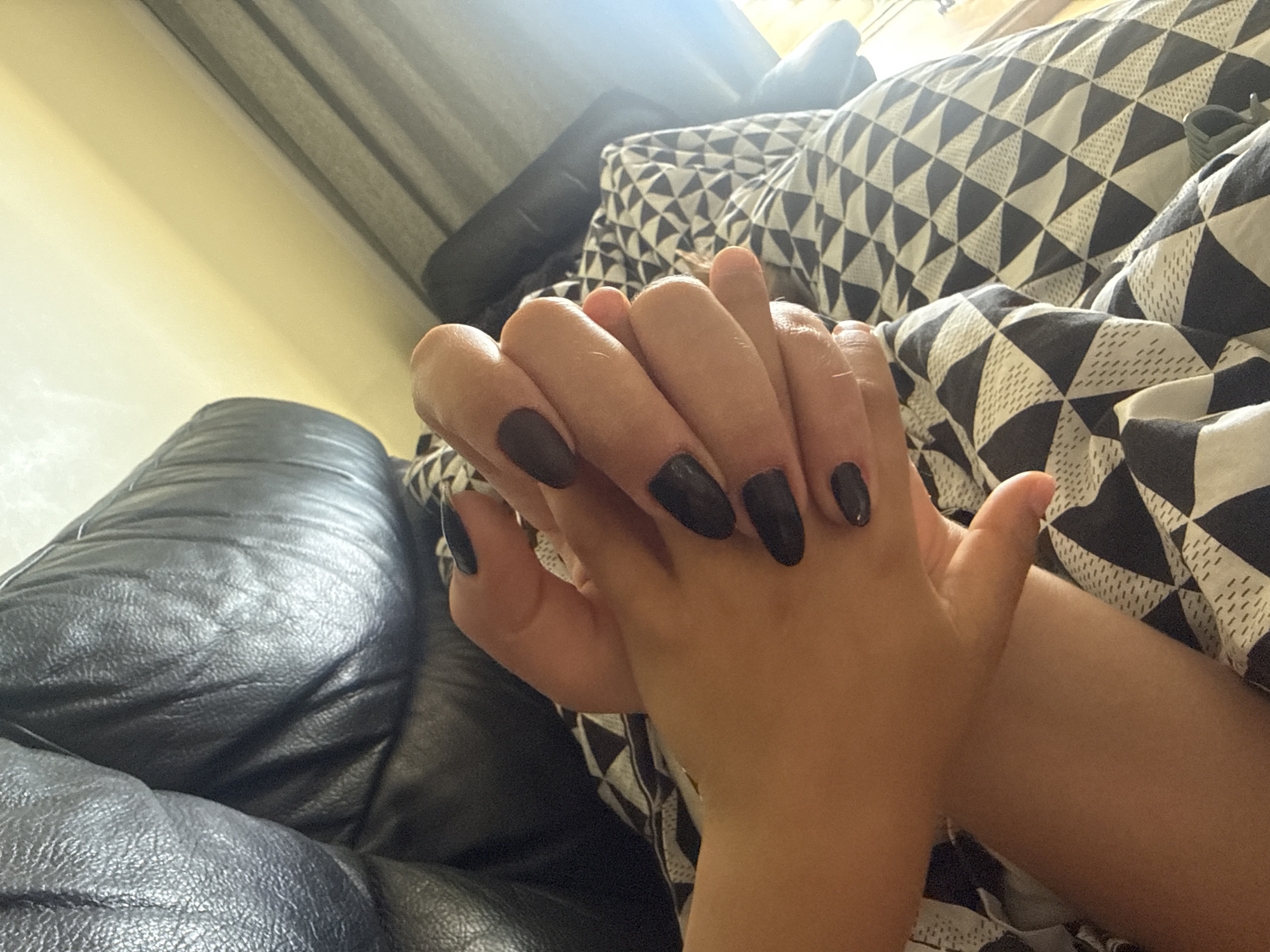 A close-up shot shows two hands with dark nail polish intertwined. The hands are resting on a black leather couch partially covered with a patterned black and white fabric. The background shows a window with a grey curtain. The location is unknown and cannot be identified as Rousset, France based on the visual information.