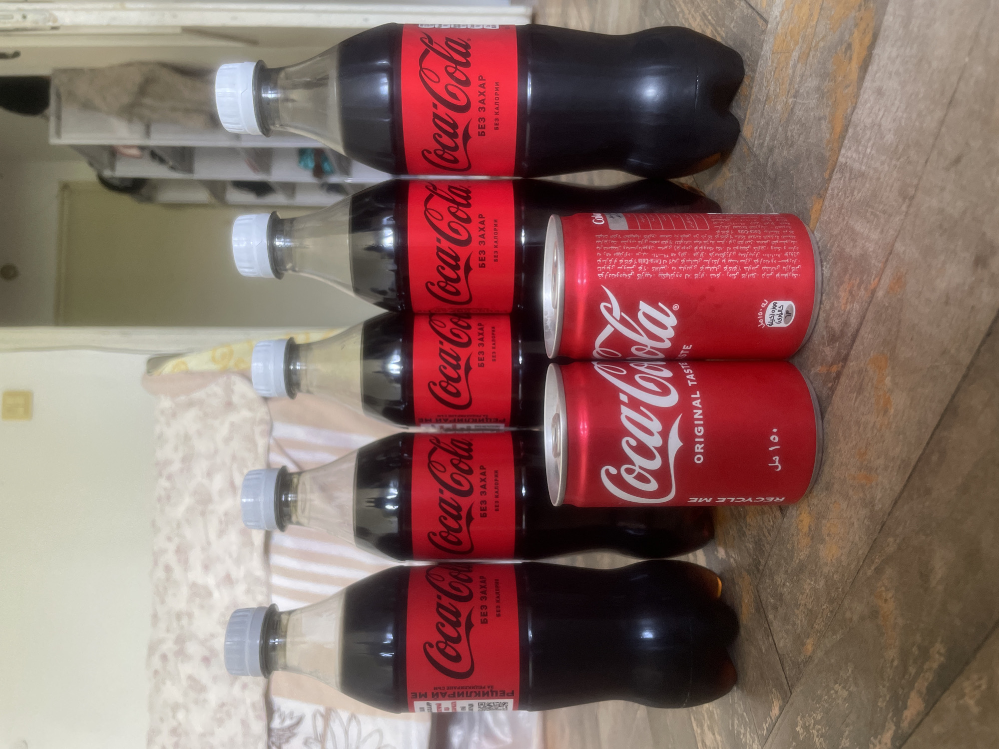 The image shows a stack of Coca-Cola products. There are five Coca-Cola bottles and two Coca-Cola cans arranged horizontally. The bottles are the 'Coca-Cola Zero Sugar' variety with text in Cyrillic. The cans are standard Coca-Cola, with some Arabic text present. The background appears to be a combination of a patterned surface and a blurred interior setting. There is no evidence suggesting it is taken in Plovdiv, Bulgaria.