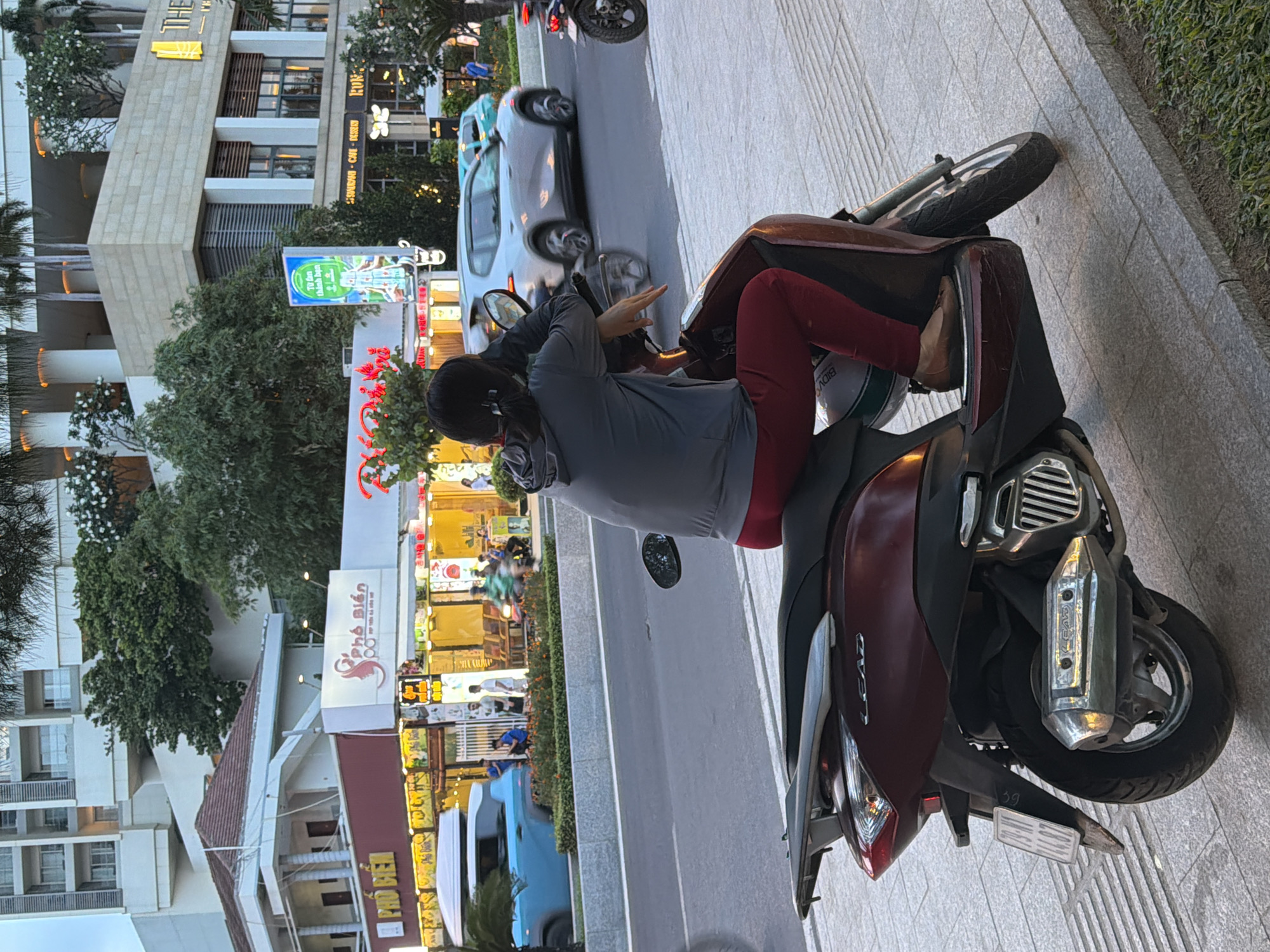 A woman wearing a grey hoodie and red pants sits on a dark red scooter, facing away from the camera. Her left arm is resting on the handlebars, and her right arm is bent, with her hand near her face. She is wearing brown flat shoes. The scooter has a license plate that reads "79-N1 293.74". The scooter is parked on a grey tiled sidewalk next to a road. In the background, there are several buildings, including a modern white building with balconies and trees on top, and a lower building with storefronts. One storefront has a sign that says "Pho Bien" in English and Vietnamese. Cars and a blue SUV are visible on the road, some in motion. Trees and greenery are present throughout the scene. The lighting suggests it is daytime, possibly late afternoon or early evening, given the warm tones. The location is identified as Nha Trang Ward, Vietnam, based on visual cues such as architecture, script on signs, and the presence of scooters.
