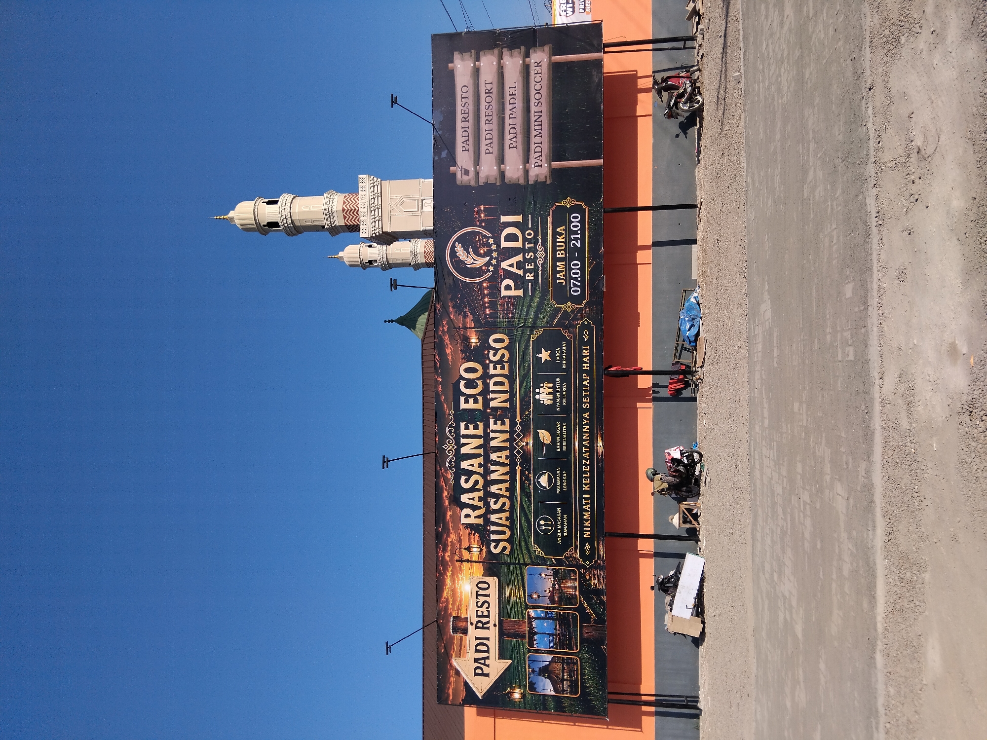 This is a landscape, eye-level, medium shot of a billboard in front of a building, under a clear blue sky. The billboard is advertising "PADI RESTO" and "SUASANANE NDESO" with images of food and facilities. It also lists "PADI RESTO", "PADI RESORT", "PADI PADEL", and "PADI MINI SOCCER" on wooden signs to the right. Two mosque minarets are visible in the background. The building behind the billboard is orange. In the foreground, there is a paved area with several motorcycles and some construction materials. This scene is likely in a rural or semi-rural area, perhaps on the outskirts of a town, given the "SUASANANE NDESO" which translates to "village atmosphere". Without further context, it is difficult to pinpoint the exact location in Jati, Indonesia.
