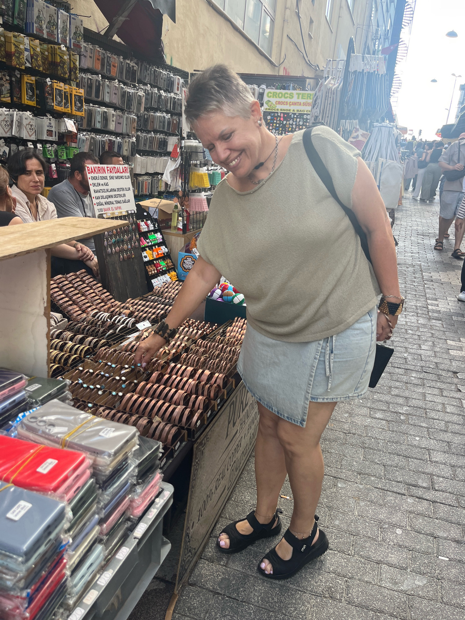 In Kadıköy, Turkey, a woman with short, gray hair is browsing a street market, examining bracelets displayed on a stall. She is wearing a light olive-green top, a light blue denim wrap skirt, and black sandals. Her right hand, adorned with a beaded bracelet, is reaching out towards the merchandise. Behind her, other vendors and shoppers are visible in the bustling marketplace, which is characterized by cobblestone pavement and open-air stalls selling various goods, including phone cases and accessories. The woman is smiling as she interacts with the market display.
