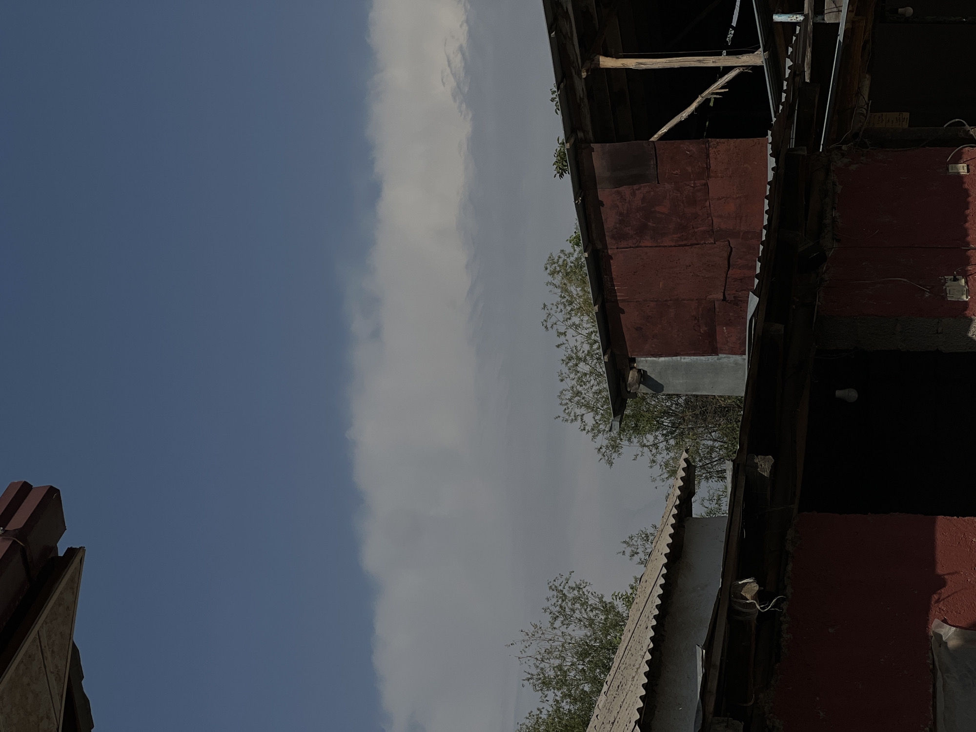 This image depicts an outdoor scene under a blue sky with some scattered clouds, featuring several structures with corrugated metal and tin roofs. In the foreground, a section of a building with a red-painted wall is visible, with exposed concrete and wiring. Above this, a corrugated metal roof extends, showing some wear and tear, with loose wires and pieces of material hanging from it. To the left, another building with a similar corrugated roof is partially visible, appearing lighter in color. Behind these structures, trees with green foliage can be seen. Further to the right, a larger structure with a rusty red tin-plated wall dominates the midground, with exposed wooden beams and a dark interior. The overall impression is one of a rural or semi-developed area. This scene is located in Karamanas, Uzbekistan.