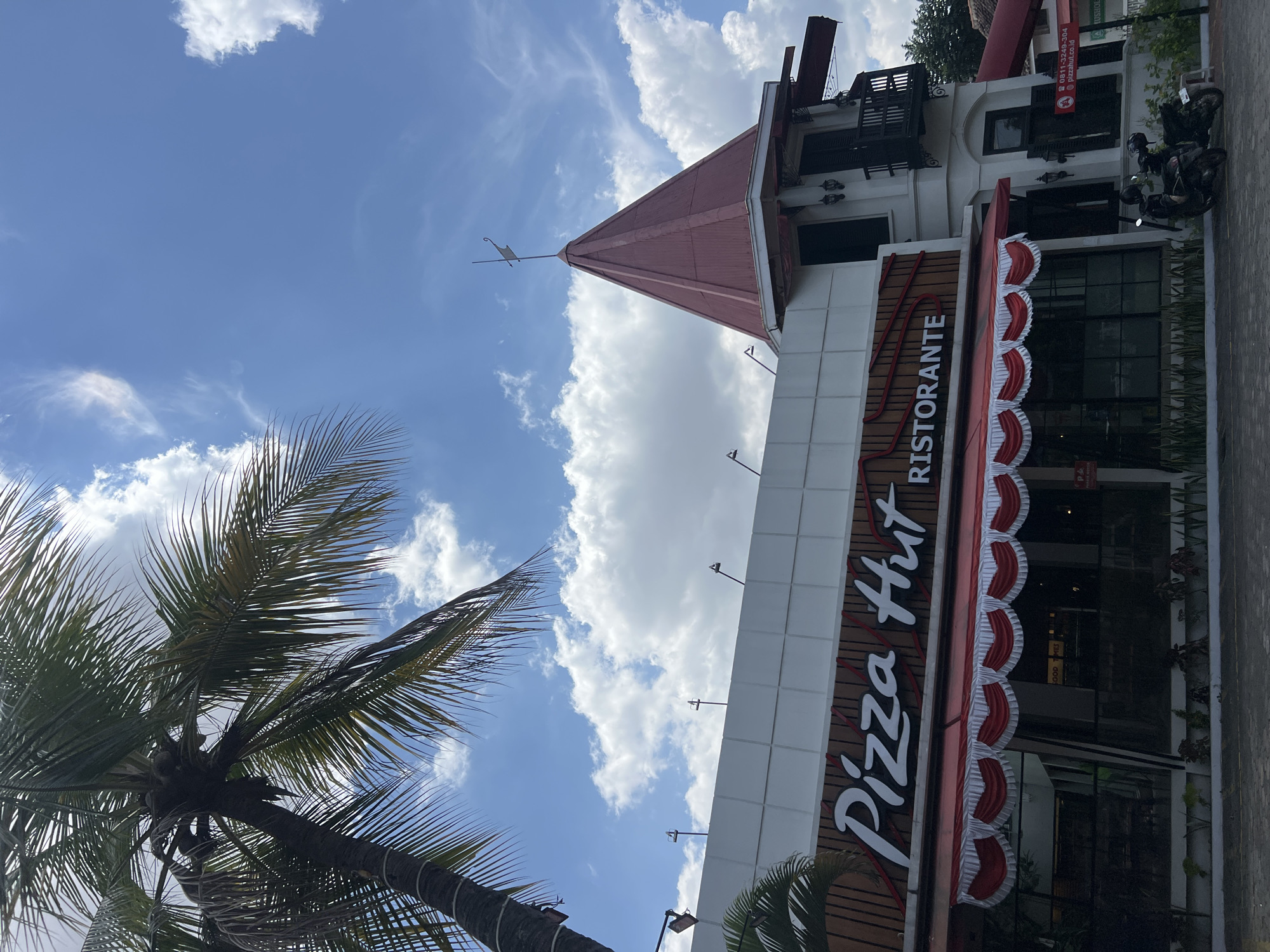 The image shows the exterior of a Pizza Hut Ristorante in the city of Yogyakarta, Indonesia. A large palm tree with coconuts is visible in the foreground to the left, with its fronds extending over the building. The building itself is modern in design, featuring a white facade with wooden accents. A prominent "Pizza Hut Ristorante" sign is displayed. A red and white awning with decorative draping runs along the front of the restaurant. To the right of the Pizza Hut, an older, white building with a conical red roof and dark shutters is attached. Two scooters are parked in front of the building. The sky above is blue with scattered white clouds. The overall scene suggests a commercial establishment in a tropical or subtropical urban environment.