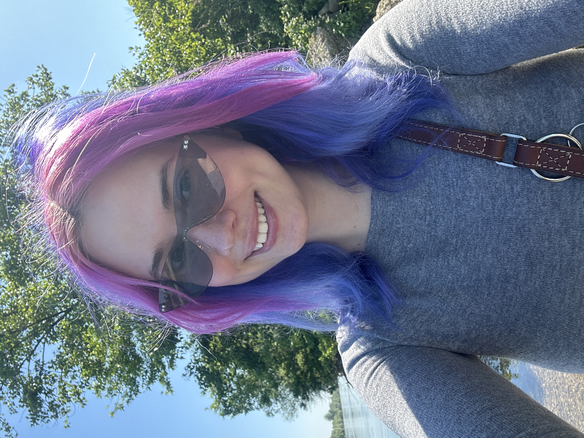 A woman with pink and purple hair is wearing sunglasses and a gray shirt. She has a brown leather leash looped over her shoulder. In the background, trees and a blue sky are visible. The location may be outdoors, near a body of water. The image does not definitively confirm it is in Zagreb, Croatia.