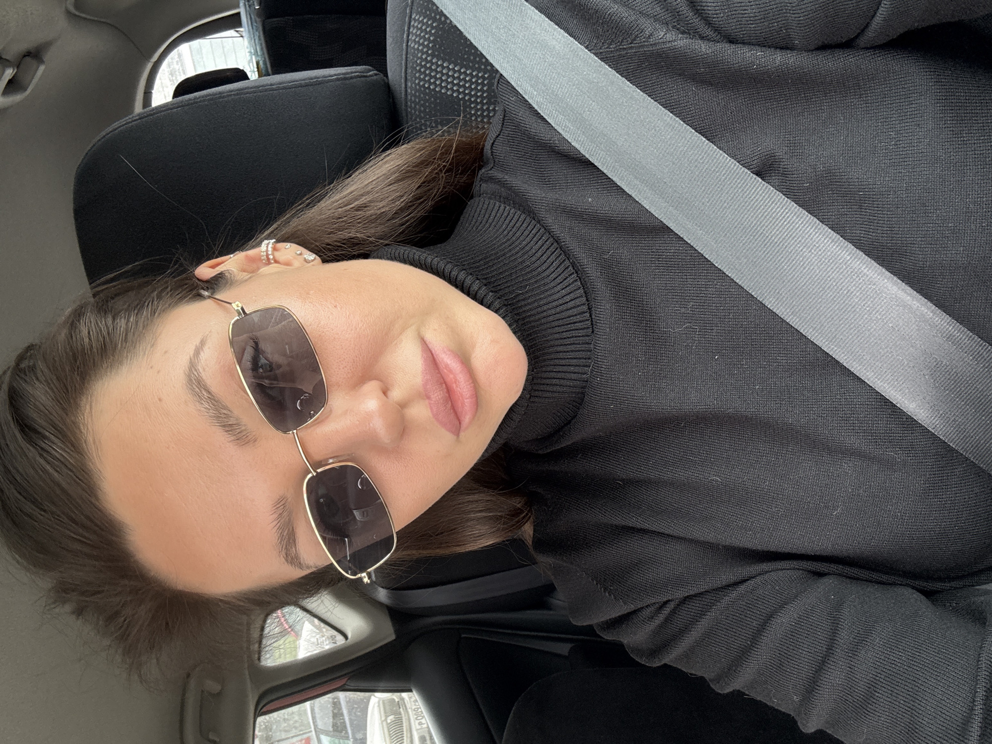 A woman wearing a black turtleneck sweater and gold-framed sunglasses is inside a car. She has a seatbelt on. She is wearing earrings on her left ear. The car is likely in Krasnoyarsk, Russia.