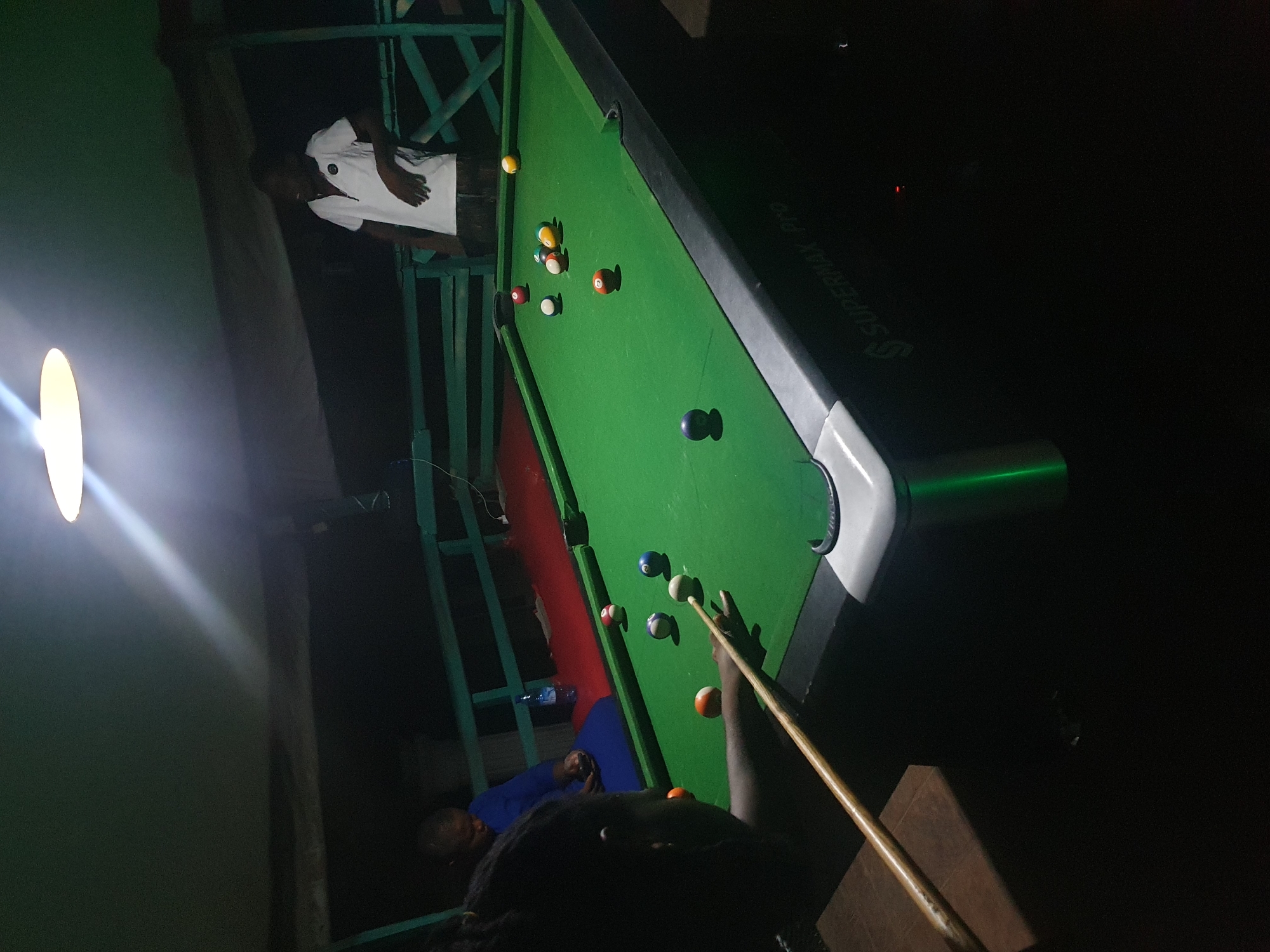 In Lokoja, Nigeria, a group of individuals are engaged in a game of billiards at what appears to be an outdoor establishment. A man in a white polo shirt is leaning on a green railing, facing away from the pool table, while another person in a blue garment sits on a bench. In the foreground, a person is actively playing pool, holding a cue stick and aiming at the balls on the green felt surface. Several billiard balls are scattered across the table, some in play and some potentially pocketed. The scene is illuminated by a bright overhead light, casting shadows and creating a dimly lit ambiance, suggestive of an evening or nighttime activity. The pool table itself has a "SUPERMAX Pro" inscription on its side.