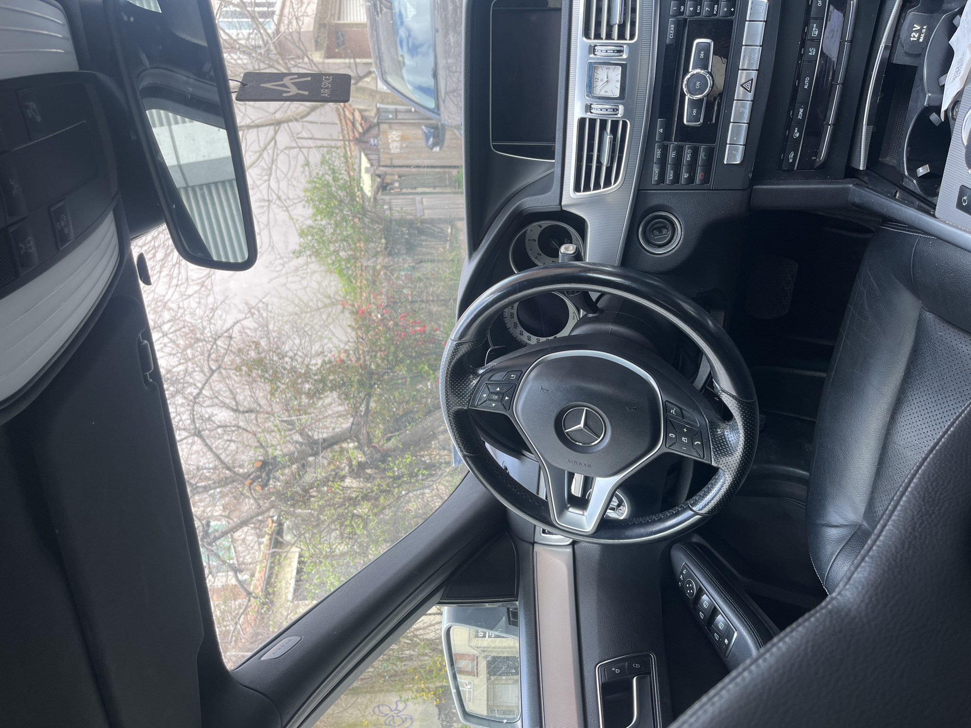 The image shows the interior of a Mercedes-Benz car. The steering wheel with the Mercedes logo is centered. The dashboard includes an analog clock, air vents, and various buttons related to the car's functions. In the rearview mirror, a street scene can be seen through the windshield, revealing a residential building. An air freshener labeled "AIR SPICE" hangs from the rearview mirror. The seats are black leather. Based on the buildings and vegetation visible, this appears to be in an urban area, possibly Varna, Bulgaria.