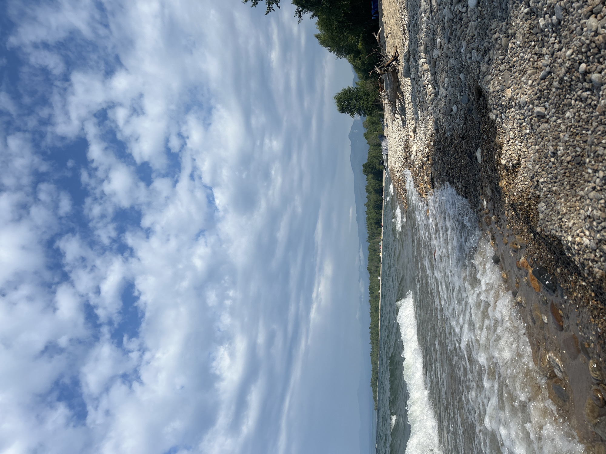 The image depicts a serene lakeside scene at Tolbazikha, Russia. The foreground is dominated by a rocky shoreline with waves gently lapping at the pebbles. The water is a deep blue-green, reflecting the overcast sky. In the distance, a dense forest lines the shore, meeting a range of misty mountains under a sky filled with scattered white clouds. A piece of driftwood rests on the beach to the right, with scattered tents and a small boat visible further down the shore. The overall impression is one of natural beauty and tranquility.