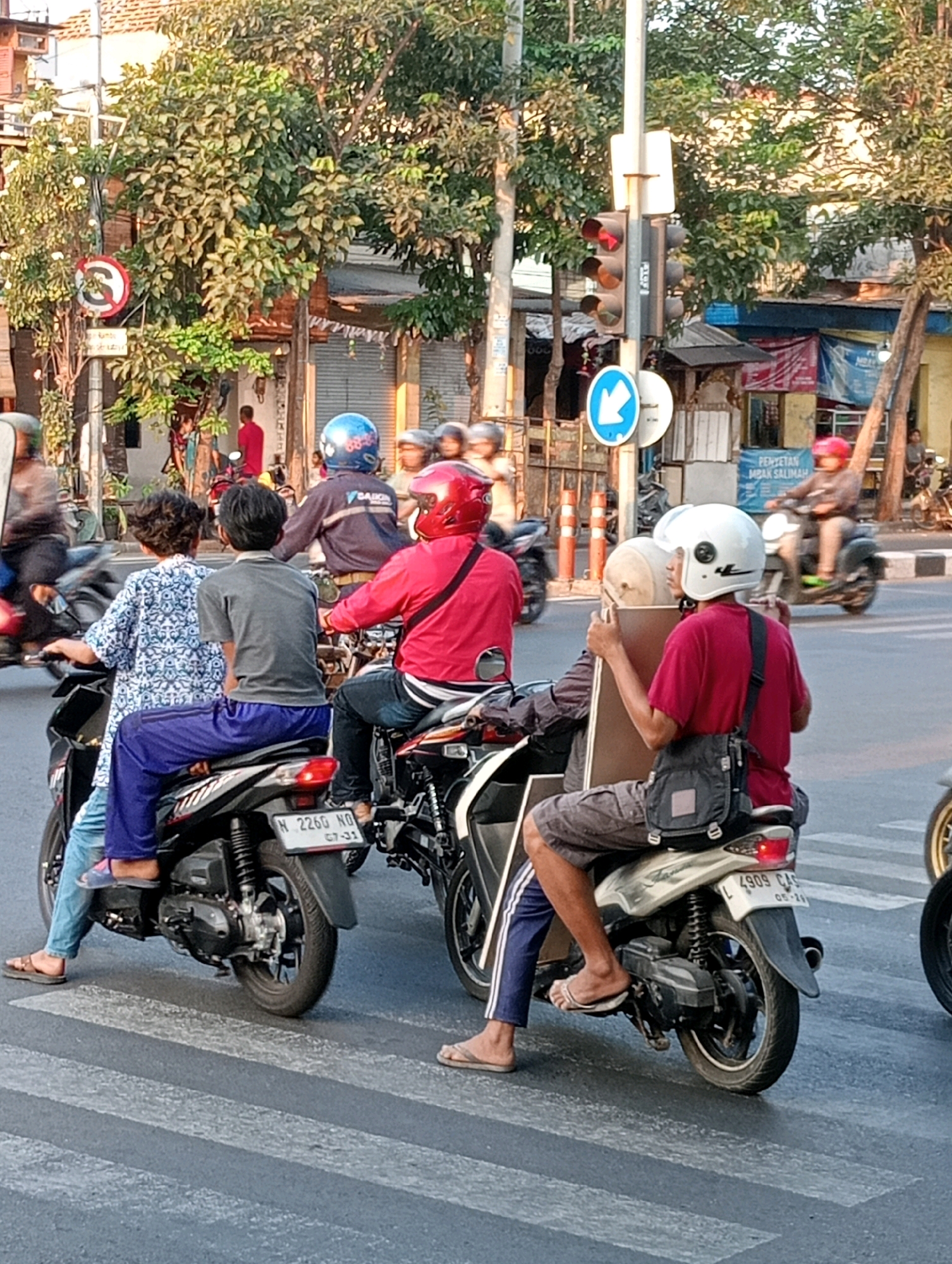 The image depicts a street scene in Surabaya, Indonesia, with several motorcycles at a crosswalk. In the foreground, two individuals are seated on a black motorcycle. The person in front, wearing a white and blue patterned shirt and blue pants, has their legs resting on the motorcycle's body. The person behind them wears a grey t-shirt and grey pants. A red motorcycle is visible to their right, with a person in a red jacket and helmet riding it. Another motorcycle, with a person wearing a red helmet and a black bag on their shoulder, is to the right of the red motorcycle. This person is holding a rectangular object. Further back, a person in a blue helmet is on a motorcycle, and other individuals are also seen on motorcycles in the background. The environment includes trees, buildings, a traffic light with a red signal illuminated, and street signs. The scene is illuminated by natural daylight, likely late afternoon given the shadows.