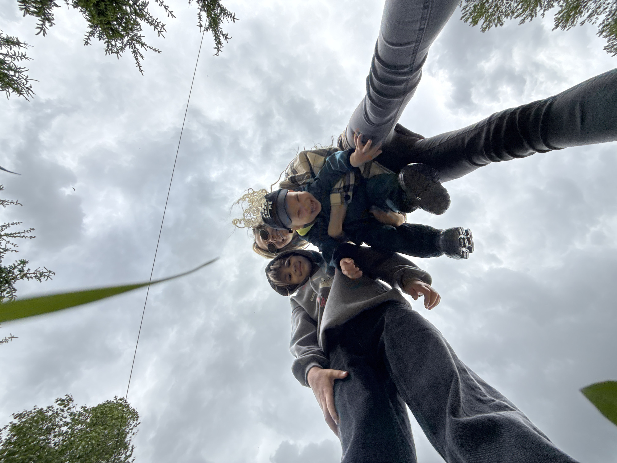 From a low angle, a woman and two children are looking down at the camera. The woman is wearing sunglasses and has blonde hair. The children appear to be male and are wearing casual clothing. The legs of another individual are seen at the top of the frame. The sky is overcast with clouds. There is vegetation in the corners of the image. The location is Unknown, Unknown.