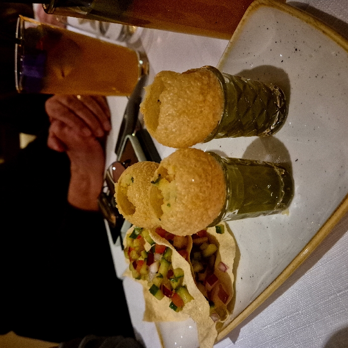 Two small glasses filled with a green liquid are topped with hollow, round, golden-brown shells. Below, two crispy, thin wafers are layered with a colorful mixture of diced vegetables, including red onions, cucumbers, and tomatoes. The arrangement sits on a square, off-white plate with a textured surface and a slightly raised golden rim. A tall glass with a brownish liquid stands in the background.
