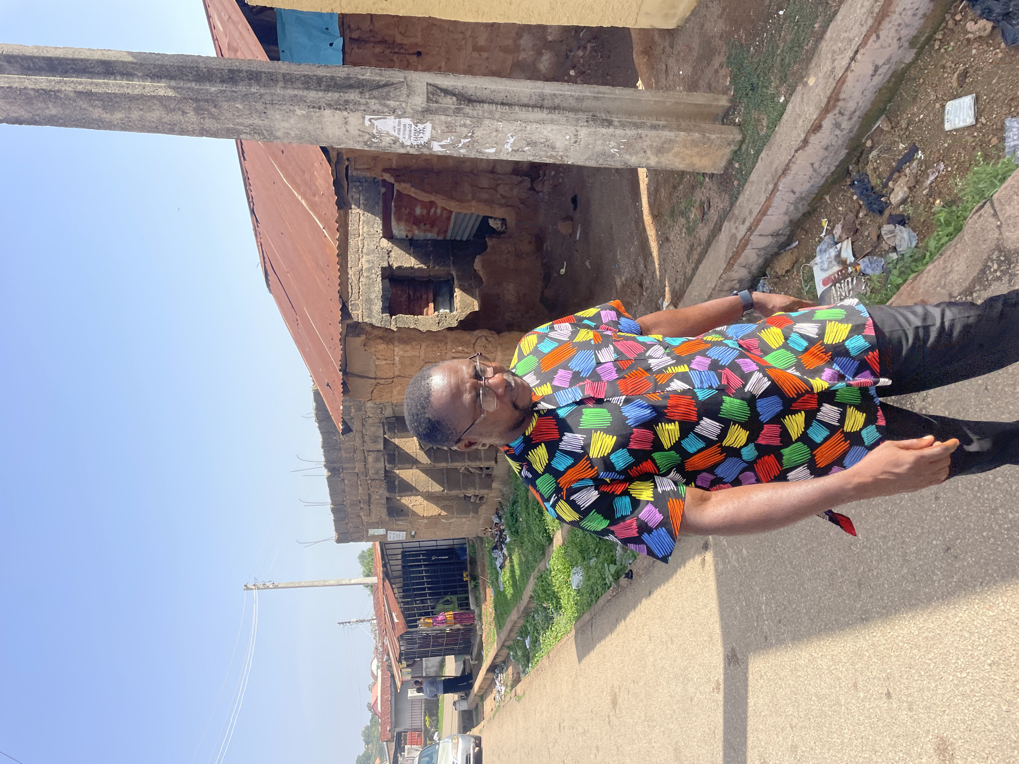 A man stands on a street in Jos, Nigeria. He is wearing glasses and a short-sleeved shirt with a black background, decorated with colorful rectangles in shades of red, blue, green, orange, yellow, and pink. He is also wearing dark pants. Behind him is a concrete building in disrepair with a corrugated metal roof. A concrete post is next to the building. The street is paved and runs alongside a concrete gutter containing trash and greenery. Further back are more buildings and vehicles. The sky is clear and blue.