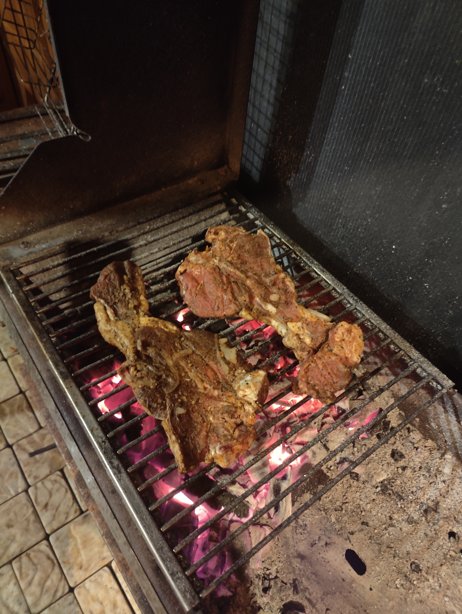 Two seasoned meat cuts, likely lamb chops due to their shape, are being grilled over glowing red embers on a metal grate. The grill appears to be in an outdoor setting, possibly a backyard or patio, with patterned stone tiles visible on the left side. The background consists of a dark, textured surface and a wire rack. The image was taken at night, indicated by the artificial lighting illuminating the scene. The location is identified as Izberbash, Russia.
