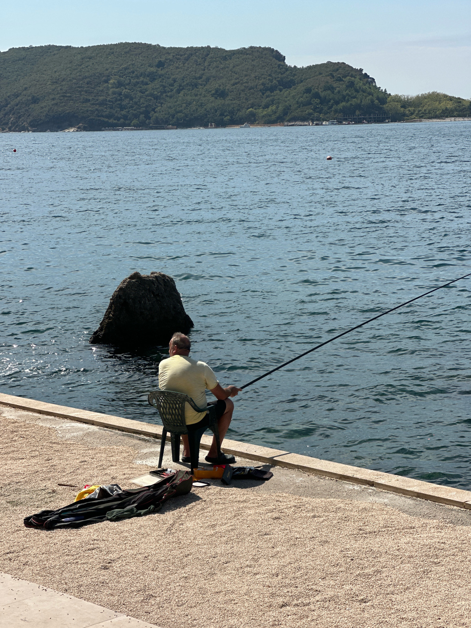 A lone man sits on a chair by the sea, facing away from the camera, holding a fishing rod. He is wearing a yellow t-shirt and dark shorts. Beside him on the ground is a fishing bag, some clothing items, and a pair of sandals. The man is positioned on a concrete walkway with a gravel-covered area behind him. The sea is calm and blue, with a large rock formation emerging from the water to his left. In the background, a tree-covered hill rises from the coastline, with a clear blue sky above. The scene appears to be located in Budva, Montenegro, given the distinctive rocky coastline and vegetation.