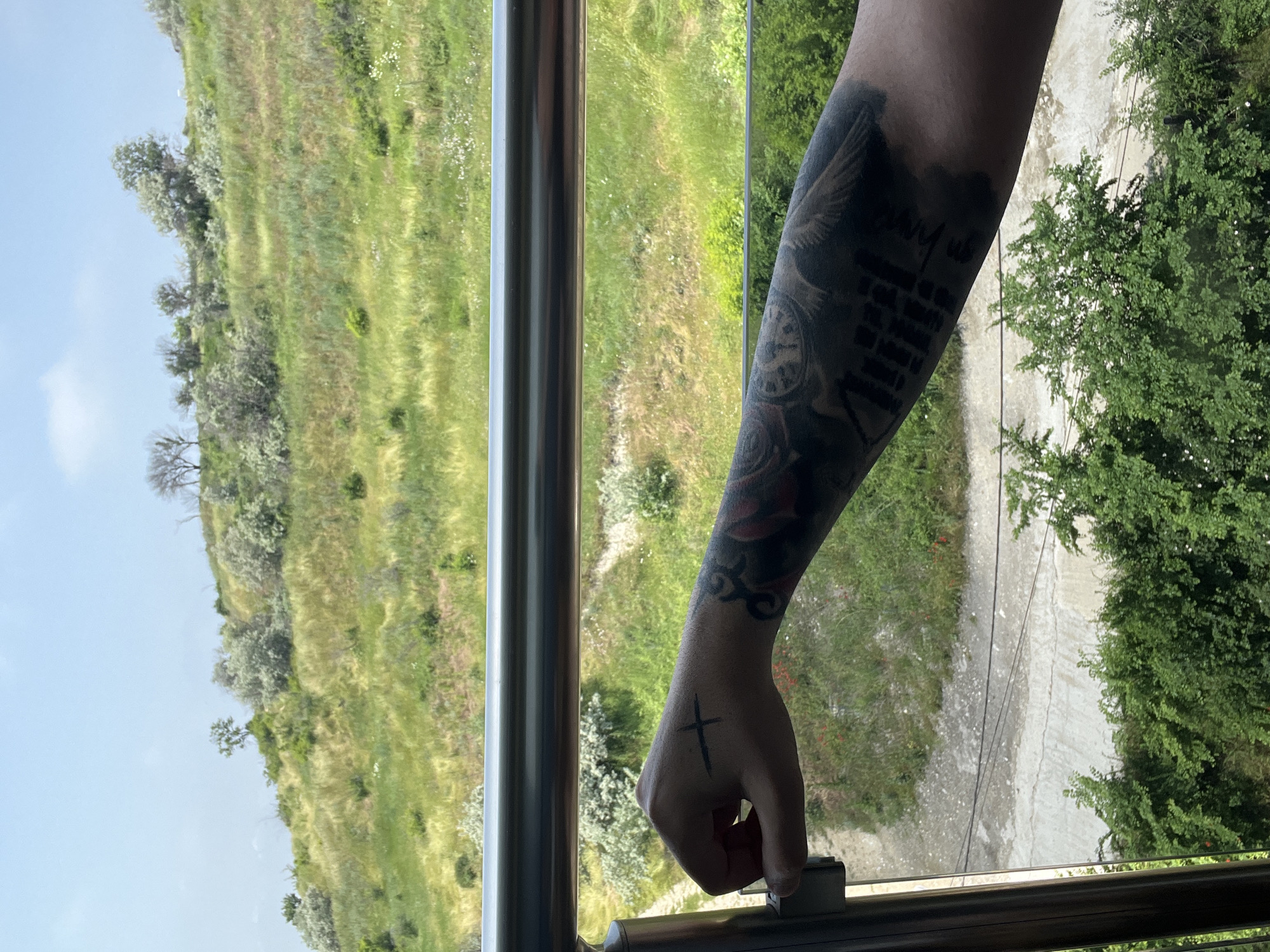 The image, originally oriented sideways, depicts a close-up view of a person's right arm and hand holding onto a metallic railing or window latch, looking out over a verdant landscape. The arm is extensively tattooed. On the forearm, there's a large, dark tattoo featuring block text that is largely illegible due to the angle and resolution, though phrases like "ONLY," "NEVER," and "WILL" are faintly discernible. Below this text, a tattoo resembling a clock face or compass is visible, and further up the arm, towards the bicep area, a design featuring a bird or wings can be seen. On the back of the hand, a simple black cross is tattooed.
The setting appears to be from a building or elevated vantage point, possibly a balcony or window. Beyond the railing, a hilly, green landscape unfolds under a clear daytime sky. The hillsides are covered in grass and scattered trees and bushes, suggesting a natural, possibly wild or semi-wild environment. A light-colored path or unpaved road is visible winding through the lower part of the landscape. The overall scene conveys a bright, sunny day. Given the provided context, the location is Varna, Bulgaria.