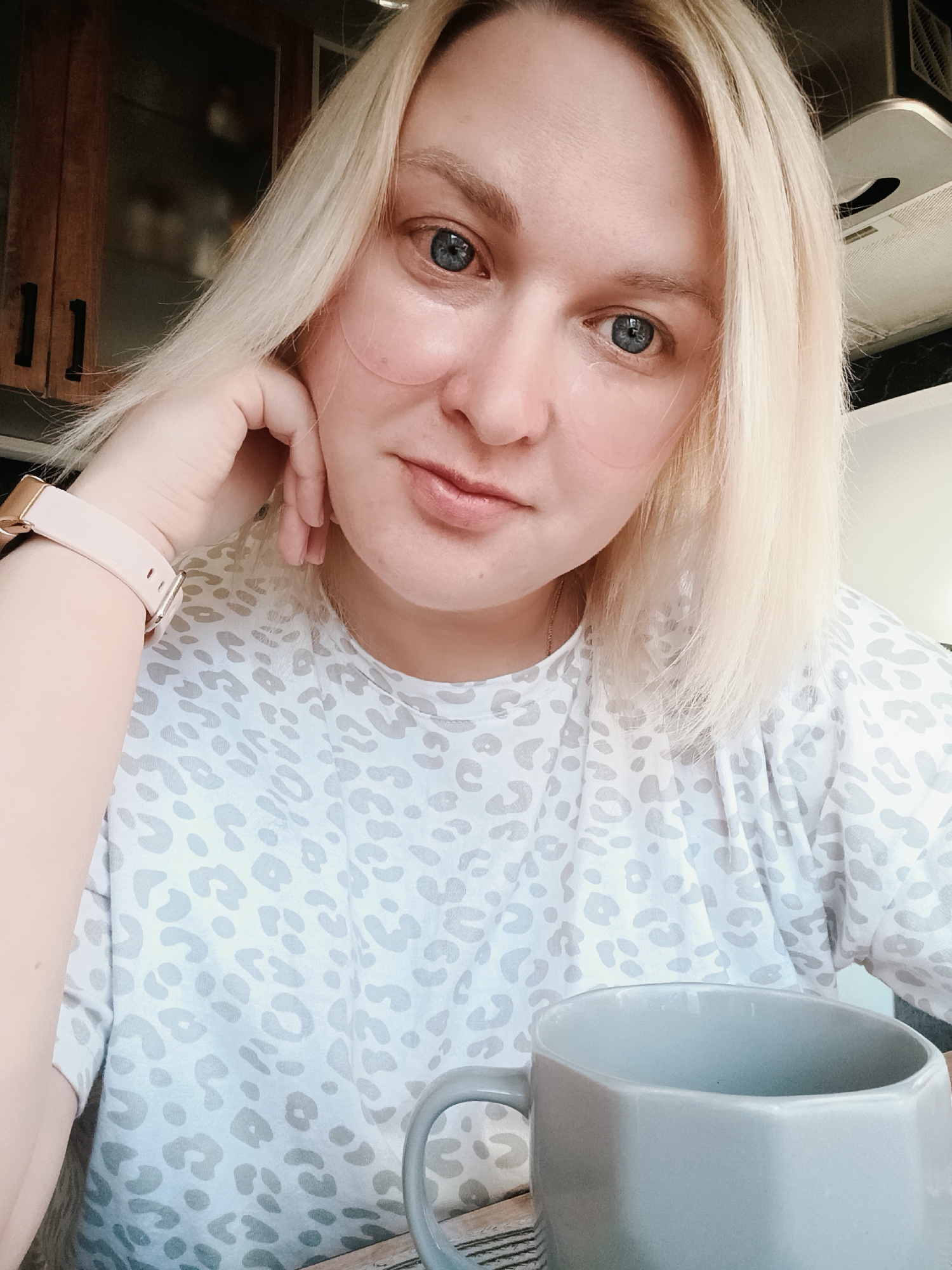 The image shows a fair-skinned woman with short, blonde hair and blue eyes. She has what appear to be cosmetic patches under her eyes. She is wearing a light-colored t-shirt with a gray leopard print pattern and a pink watch on her left wrist. She is resting her head on her hand. A gray mug is visible in the foreground, partially obscuring her shirt. The background includes a wooden cabinet and a kitchen vent. The photograph was taken in Chaikovsky, Russia.