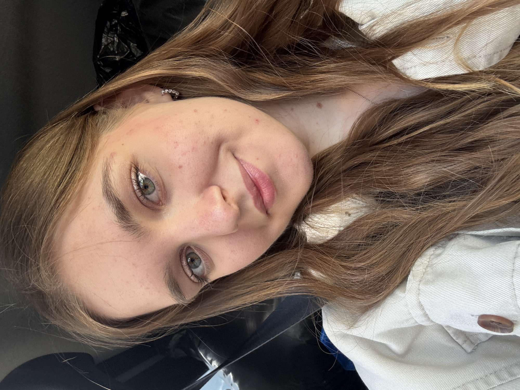 A young woman with light brown, wavy hair poses indoors. She has fair skin, light eyes, and wears a white denim jacket. An earring is visible in her left ear. A dark, possibly black, object is behind her head. The location in Perm, Russia is not discernible from the image.