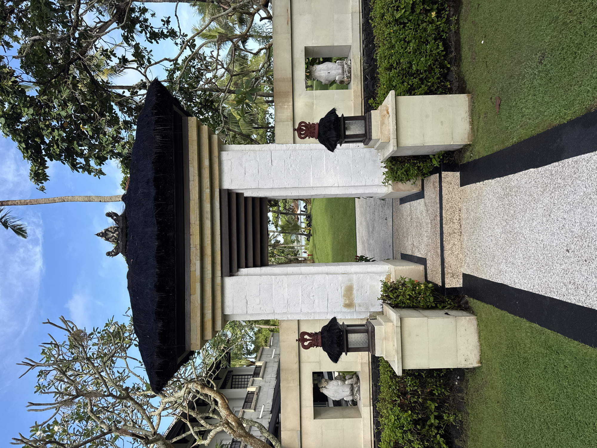 The image displays a traditional Balinese-style entrance gate under a bright blue sky, set in an outdoor, lush environment. The gate, constructed from light-colored stone blocks, features two substantial pillars supporting a multi-layered, dark thatched roof. Decorative elements include ornate stone carvings at the top of the gate structure and two black lanterns with a bronze crown-like design positioned on stone pedestals flanking the inner side of the archway.
Through the archway, a two-toned pathway leads into a manicured green lawn. The path consists of a light-colored, speckled surface bordered by a dark, possibly asphalt, strip. Beyond the gate and lawn, more trees and hints of buildings are visible in the distance. On either side of the pathway, within the stone gate structure, are recessed niches, each containing a white stone statue, possibly of a deity like Ganesha or a similar figure, enhancing the cultural aesthetic.
The surroundings are verdant, with dense green bushes lining the base of the gate and various trees, including tall palm trees and other leafy varieties, framing the scene against the clear, blue sky with scattered white clouds. The bright, even lighting suggests it is daytime, likely morning or early afternoon. The overall scene conveys a sense of peace and cultural authenticity, characteristic of a tropical resort or temple entrance in Kuta Selatan, Indonesia. There is no visible text in the image.