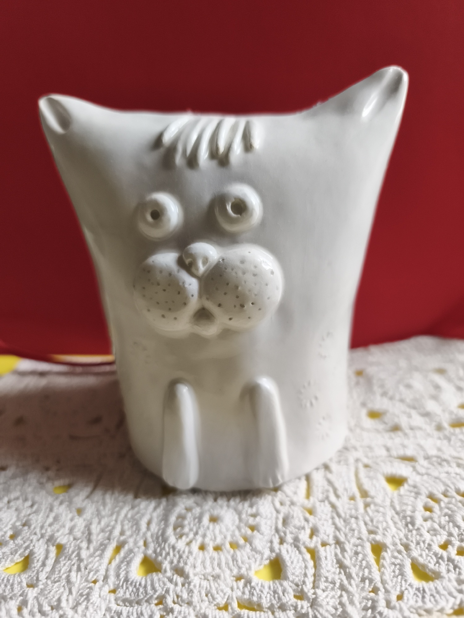 A white ceramic cat figurine sits on a white crocheted doily, which is placed on a yellow surface. The figurine has a stylized face with large, round eyes, a prominent nose with textured cheeks, and short, stubby legs. The background is a plain red surface. The scene is indoors, with no indication of location within the city of Saint-Denis, France. There are no people or actions depicted in the image.