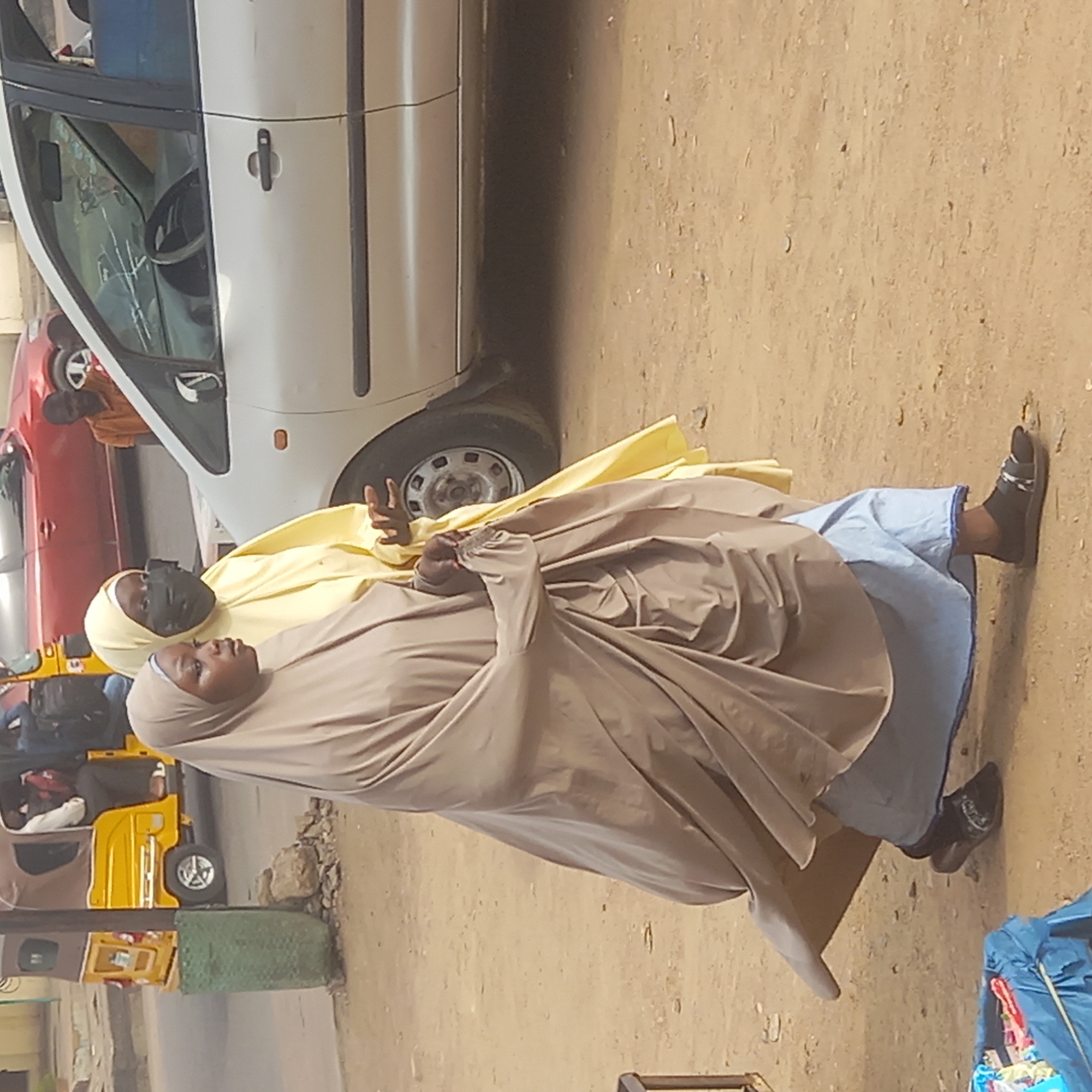 Two women are featured prominently in this outdoor street scene, captured in what appears to be Dadi Riba, Nigeria. The woman in the foreground is dressed in a light brown or taupe full-body covering (hijab or abaya) and a matching head covering, with her face fully visible and looking towards the left. Behind her, another woman wears a bright yellow full-body covering and a matching head covering, with a dark face covering (possibly a niqab) exposing only her eyes. This second woman is raising her right hand, displaying a 'peace' or 'victory' sign.
They are standing on an unpaved, sandy-colored ground that takes up a significant portion of the image. Various vehicles are parked in the background. A white car, possibly a minivan, is visible on the right, with its front wheel and part of its interior (including a steering wheel) seen through the window. Behind it, a red car is partially visible. Further to the left, a yellow motorized tricycle taxi, commonly known as a "keke napep," is parked. A green, low barrier or planter is situated near the keke. To the right of the women's feet, a blue bag or tarp with contents is resting on the ground.
The scene appears to be taking place during daylight hours, indicated by the natural and even lighting. There are no clear indications of specific weather conditions other than brightness. No discernible text is visible anywhere in the image.