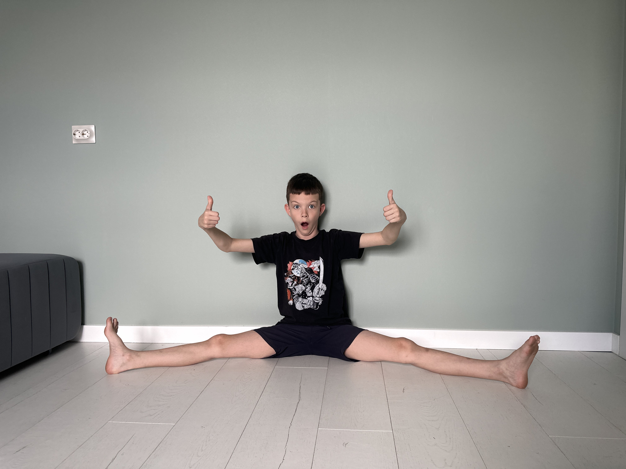 A fair-skinned boy in a black t-shirt and dark shorts is sitting on a light-colored wooden floor in a full split, with his arms outstretched and thumbs up. His expression is one of surprise or excitement, with his mouth open. Behind him is a light green wall with a double electrical outlet on the left. Part of a gray couch is visible in the lower-left corner of the frame. The room is well-lit, suggesting a modern, residential interior. The location is Прикамский, Russia.