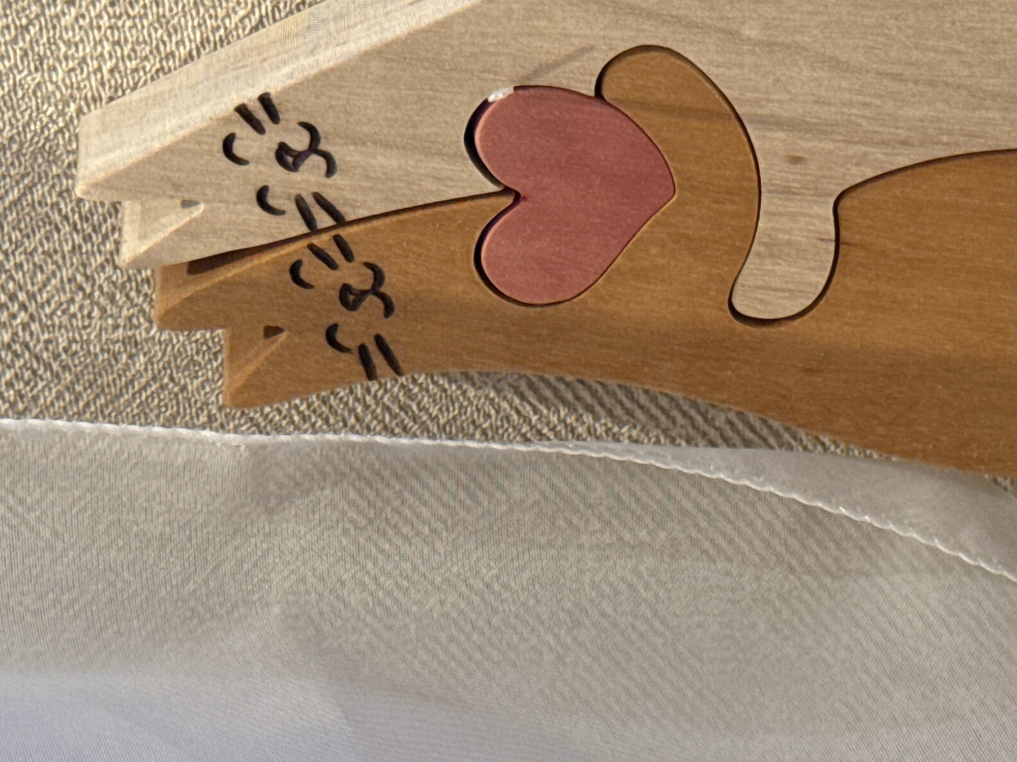A close-up, high-angle shot depicts two interlocking wooden cat puzzle pieces resting on textured fabric. The upper cat piece is made of lighter-toned wood, while the lower piece is crafted from darker, reddish-brown wood. Both cats feature minimalist black markings representing eyes and whiskers. A small, reddish-pink heart-shaped piece is snugly fitted between the bodies of the two cats, forming part of their interlocking design. The wood grain is visible on all pieces.
Below the wooden puzzle, there is a clear line of stitching separating a textured, light beige fabric in the background from a smoother, off-white fabric in the foreground. The foreground fabric displays a subtle herringbone weave pattern. The background fabric has a rougher, more irregular texture. The lighting is soft, suggesting an indoor setting. There are no people visible, and no discernible activity other than the static arrangement of the puzzle pieces. No text is visible in the image. No specific geographical or time-of-day cues are present.