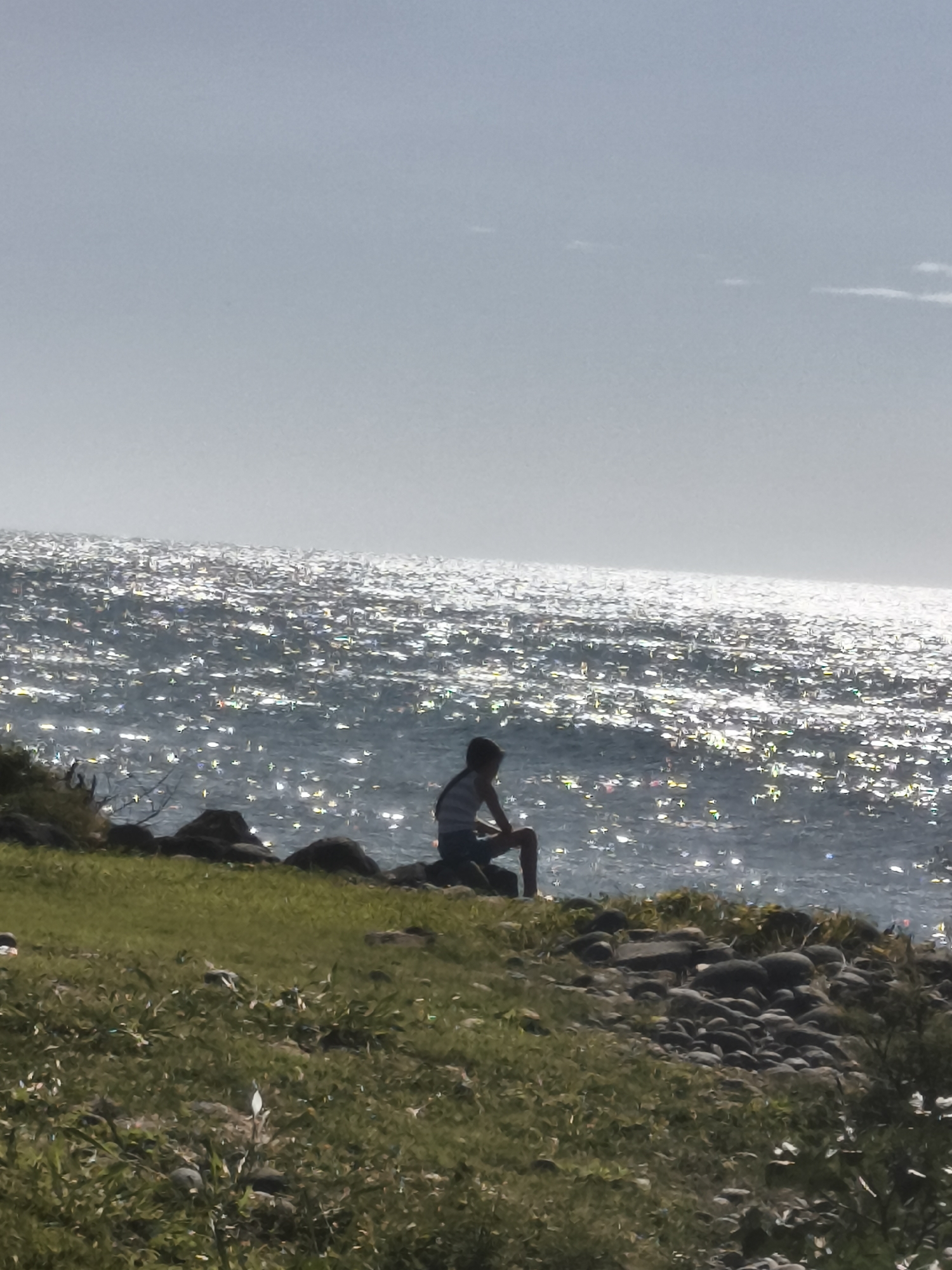 The image shows a silhouetted person sitting on rocks by the sea. The sea is glittering with sunlight, and the sky is overcast. The person is wearing a striped shirt and shorts. The foreground is a grassy embankment with scattered rocks. The location is not identifiable as Sainte-Marie, France, as there are no specific landmarks or contextual clues to indicate this.