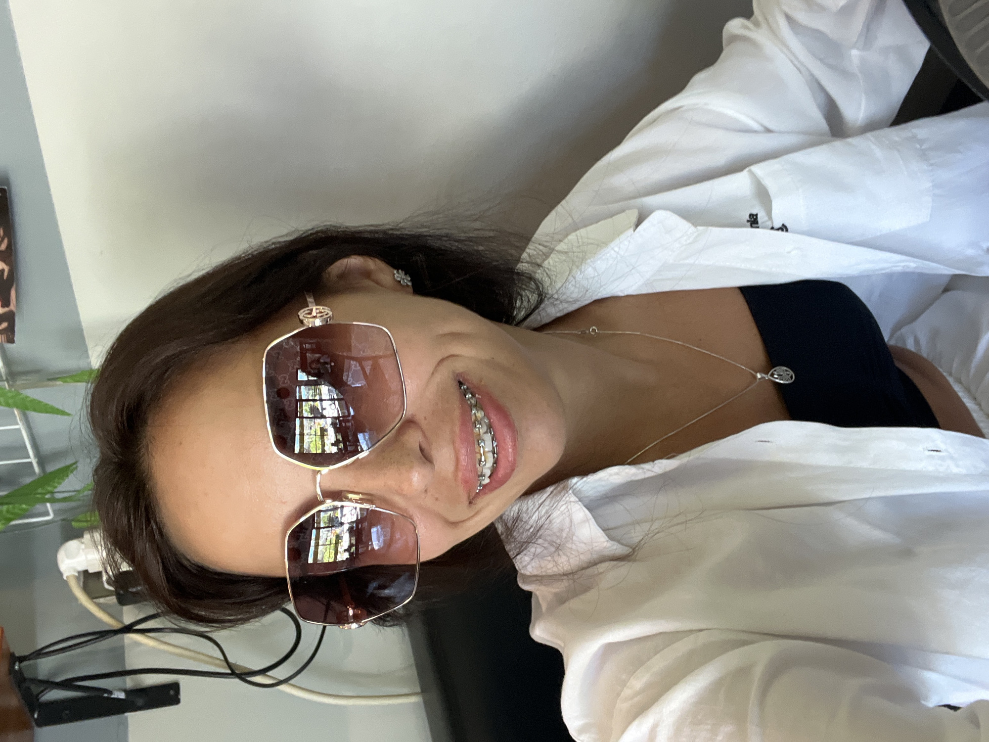 A woman with dark hair and braces smiles while wearing sunglasses, a white collared shirt over a black top, and a silver necklace. The reflection in her sunglasses shows buildings and greenery, suggesting an urban environment. The presence of electrical cords and a metal rack points to an indoor setting, possibly a cafe or office. There is no definitive information to confirm a location in Kuta Selatan, Indonesia.