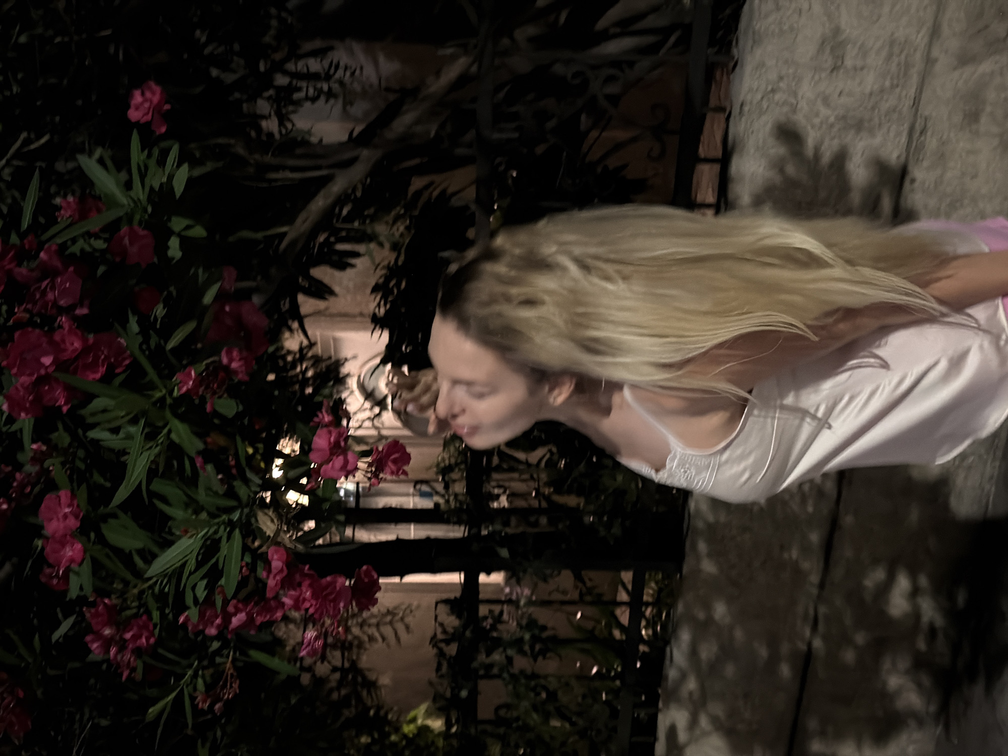 In Budva, Montenegro, at night, a young woman with long blonde hair is seen from the chest up, looking upwards with her eyes closed, surrounded by a tree with pink flowers. She is wearing a white satin slip with lace detailing on the straps. The background shows a building with illuminated windows and a dark fence. The lighting is dim, creating shadows on a textured wall in the foreground.