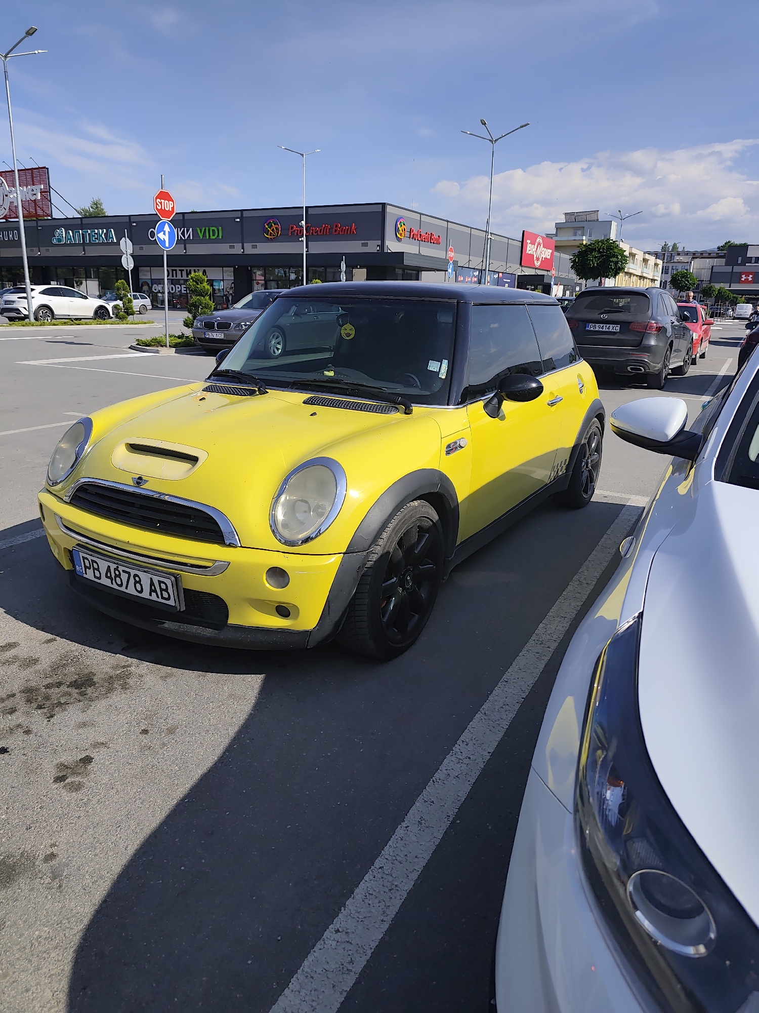 The image shows a yellow Mini Cooper with a black roof parked in a parking lot. The car has a Bulgarian license plate, "PB 4878 AB". Other cars are parked nearby, including a grey SUV (license plate "PB 9414 XC") and a white vehicle. In the background are several stores, including an "АПТЕКА", "СКИ VIDI", "ProCredit Bank" and a "Tехно Mаrкет" (Tech Market). A stop sign is also visible. The location is in Plovdiv, Bulgaria. The image was taken during daylight hours.