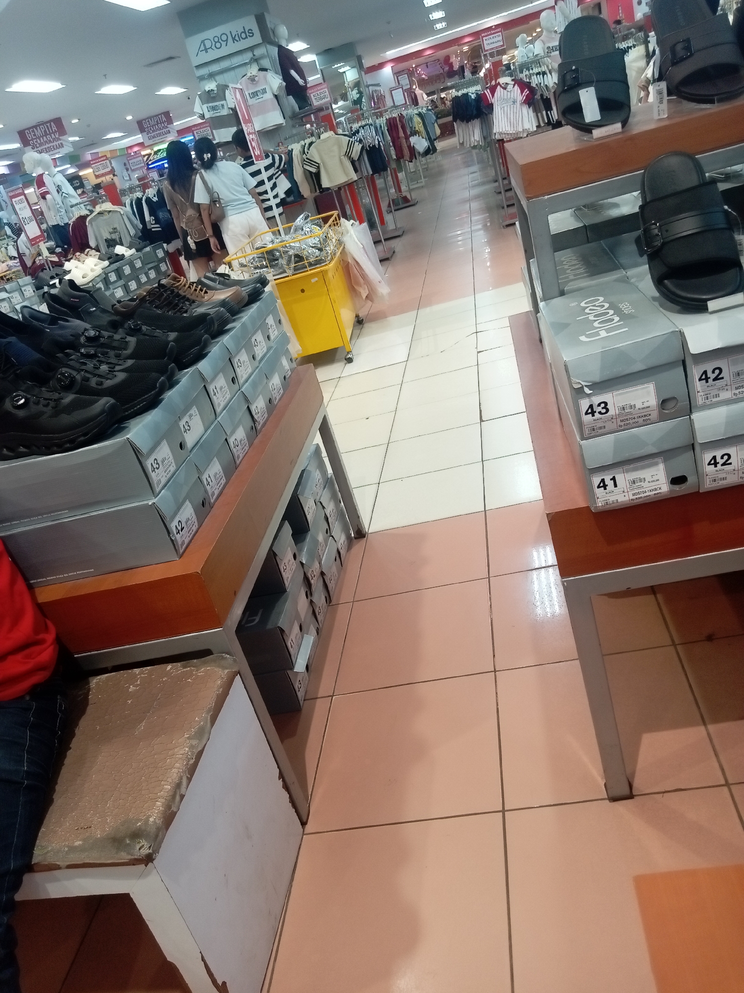 The image captures an indoor scene within a retail store, likely a shoe store or a department store with a shoe section, in Batam, Indonesia. The environment appears to be a well-lit commercial space with tiled flooring and rows of shelves displaying merchandise. In the foreground, there are stacks of shoe boxes, some labeled with sizes like "43", "41", and "42", and brands like "Fodee". On top of some shelves, there are display shoes, including black sandals.
Further into the store, clothing racks are visible, with various garments, including shirts and what appear to be children's clothing, some with signs indicating brands like "AR89 kids". Several individuals are present in the scene. Two women are standing and looking at the merchandise. One is wearing a light-colored top and white shorts, and the other is wearing a dark top and has a bag slung over her shoulder. Another person, partially visible in a red top and jeans, is seated on a bench or stool in the lower left portion of the image. A yellow shopping cart is also present in the mid-ground. The overall impression is a typical retail environment with customers browsing and shopping.