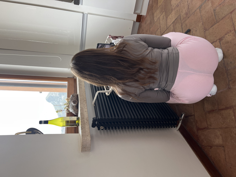 The image displays an indoor scene within the city of Cava de' Tirreni, Italy. A female subject is positioned centrally, viewed from the rear, in a crouched or squatting posture. She has long, brown, wavy hair. Her attire consists of a long-sleeved grey top, light pink form-fitting pants, and white socks or slippers. Her attention is directed towards a mobile device, likely a smartphone, which is mounted on a small white metal stand placed directly on the floor in front of her.
To the left of the subject, a black, horizontally ribbed radiator is affixed to a white wall. The flooring consists of terracotta-colored tiles. Behind the subject, a window with a light brown wooden frame provides a view of an outdoor environment featuring a prominent green mountain peak under a bright sky. On the stone windowsill, a partially filled clear green glass bottle, identified by a "Lagaria" label, stands next to a small succulent plant potted in a rustic, log-shaped planter. To the right of the subject, white, flat-paneled cabinetry or appliances are visible along a wall, with a thin silver chain or cord hanging vertically.