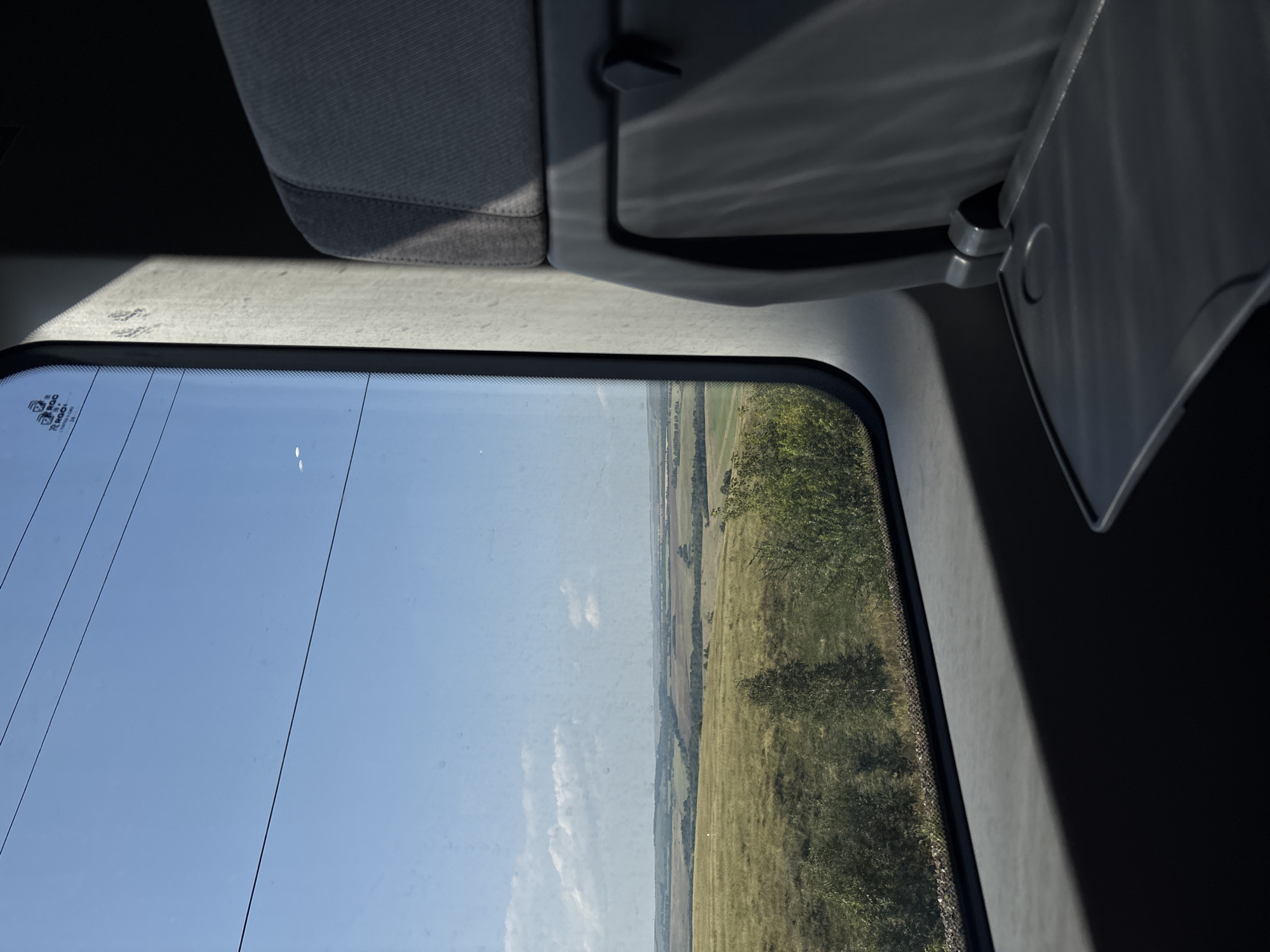 The image depicts a view from inside a train looking out the window at a rural landscape under a clear blue sky. The foreground shows the interior of a train carriage with a gray fabric seat and a fold-down tray table. Sunlight casts a shadow across the seat and tray table. The window is slightly dirty and reflects some of the interior. Outside the window, a vast expanse of rolling hills and fields unfolds, dotted with trees and agricultural land. The fields are a mixture of green and golden hues, suggesting a summer or early autumn setting. The sky is a bright, cloudless blue, with a few faint white clouds visible in the distance. There are power lines visible across the sky, indicating the train is likely on an active route. There are no people visible in the image. The location in the city of Shafranovo, Russia is not discernible from the image itself.
