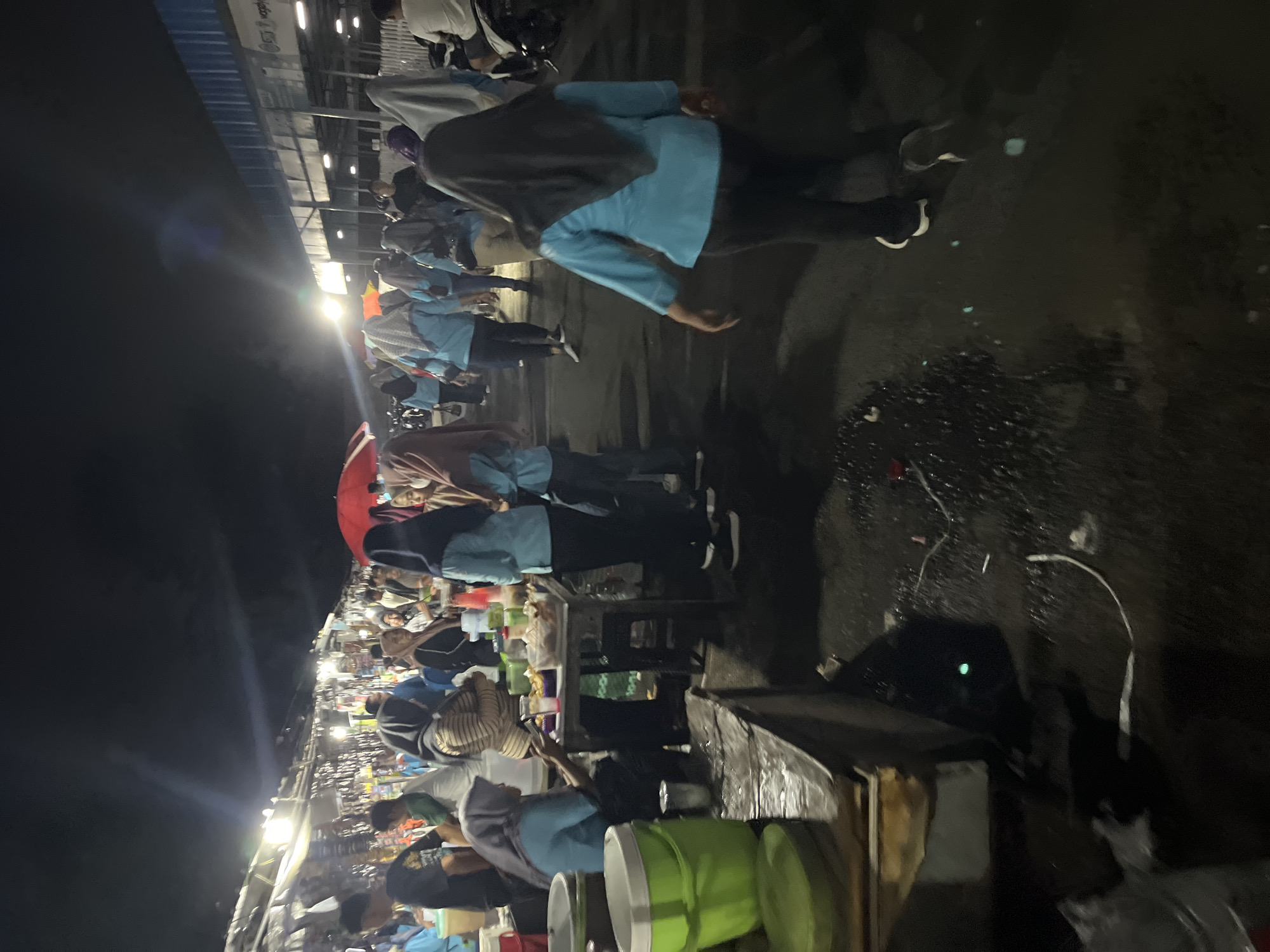 The image depicts a night scene at a market in Pelang, Indonesia. A group of people, predominantly women wearing headscarves and light blue tops or tunics, are walking through a wet, dimly lit outdoor area. Some individuals are interacting with vendors at stalls lined with merchandise. A red umbrella is visible among the crowd. In the foreground, there are large green buckets and a wooden table laden with food containers. The ground appears to be asphalt and is wet, reflecting the surrounding lights, suggesting recent rain or cleaning. The market stalls feature various goods, and some have overhead lighting illuminating the displays. The overall atmosphere suggests a busy local market operating after dark.